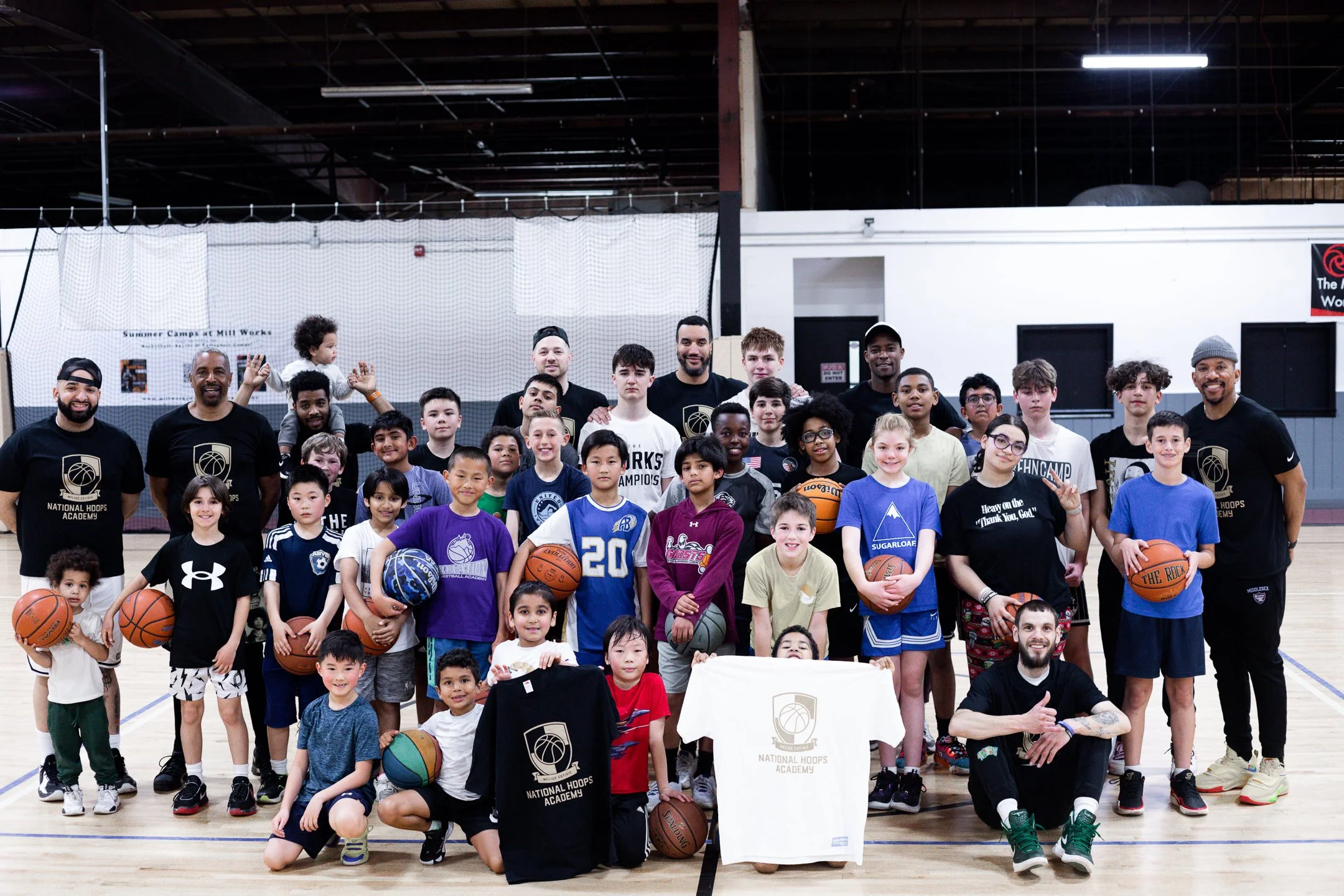Group of children and coaches posing for a photo on an indoor basketball court, some holding basketballs, at a summer camp at Mill Works.