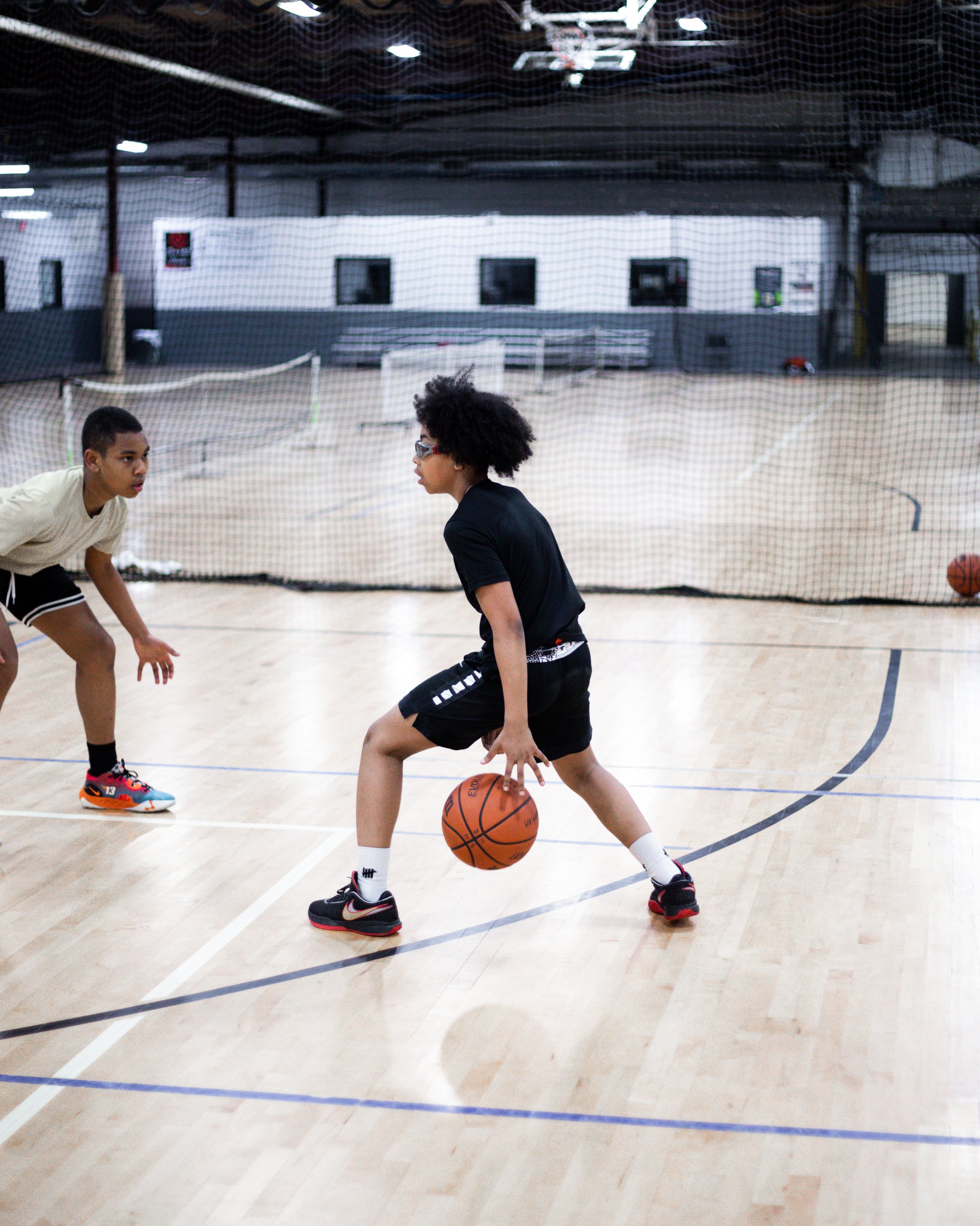 Two children playing basketball in an indoor gymnasium, with one child dribbling the ball and the other in a defensive stance.