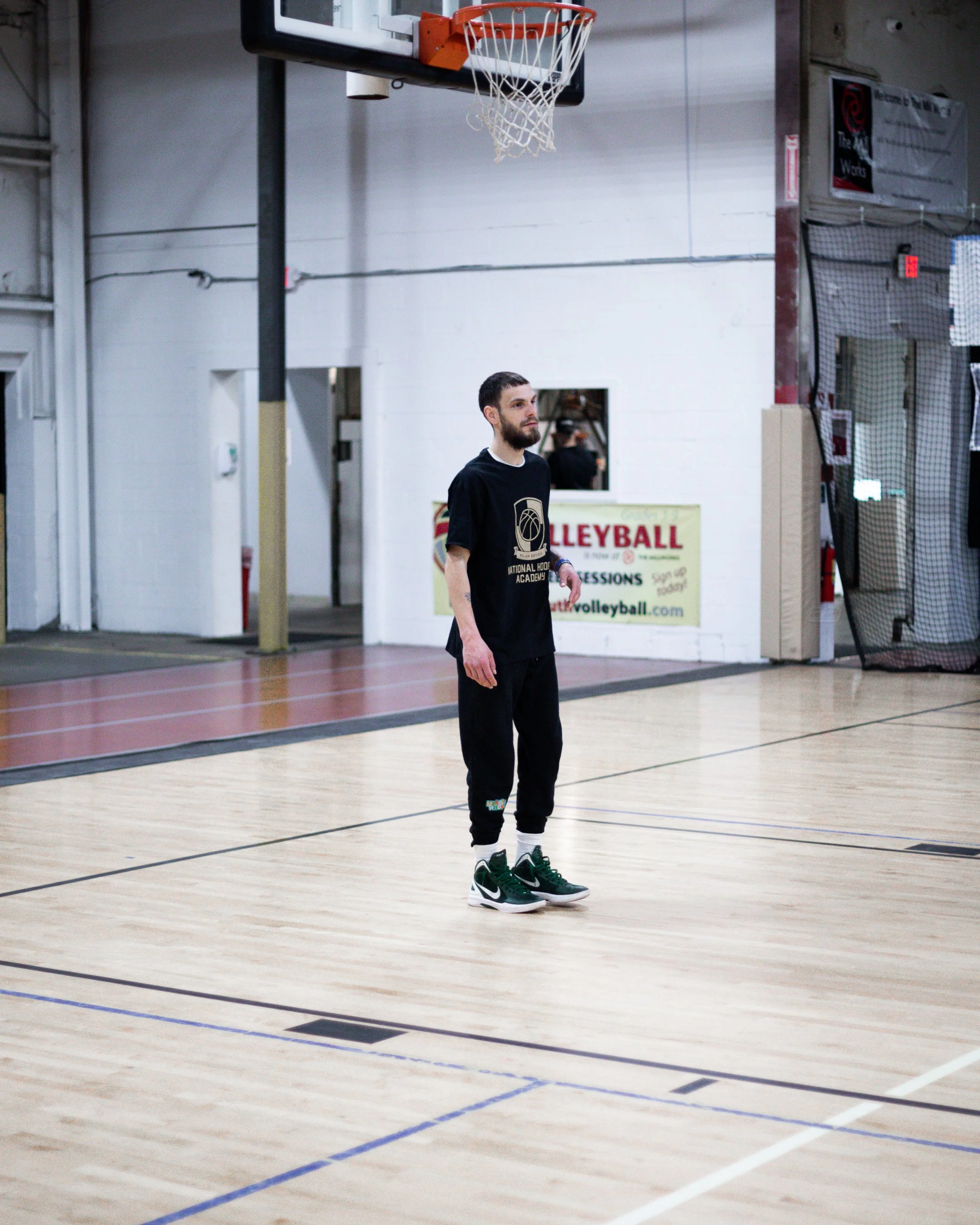 A man standing alone on an indoor basketball court, wearing black athletic clothes and green sneakers, under a basketball hoop.