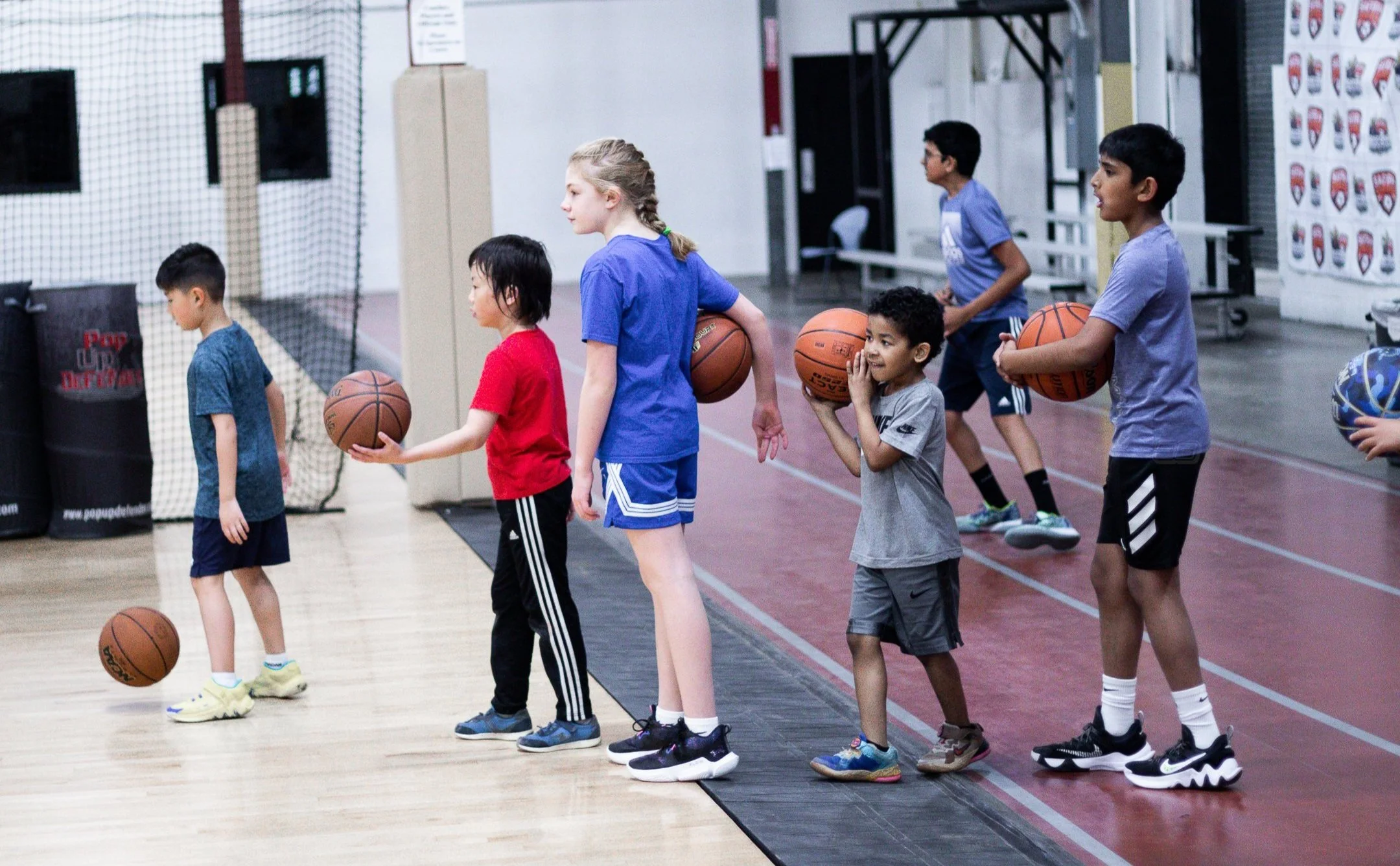 A group of children practicing basketball in a gym, each holding a basketball and standing in a line.