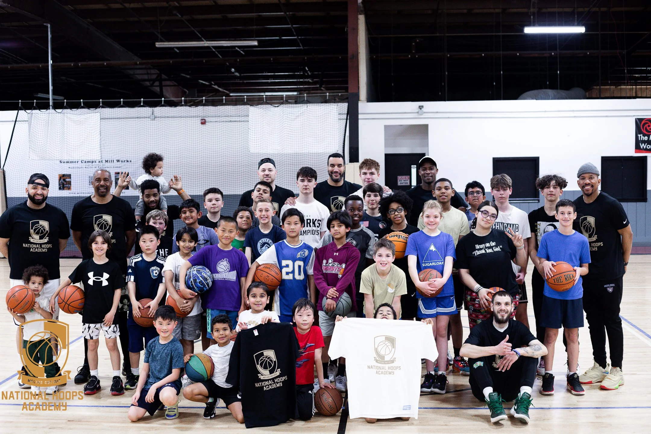 Group photo of children and coaches at a basketball camp in an indoor gym, with some children holding basketballs and displaying T-shirts with the 'National Hoops Academy' logo.