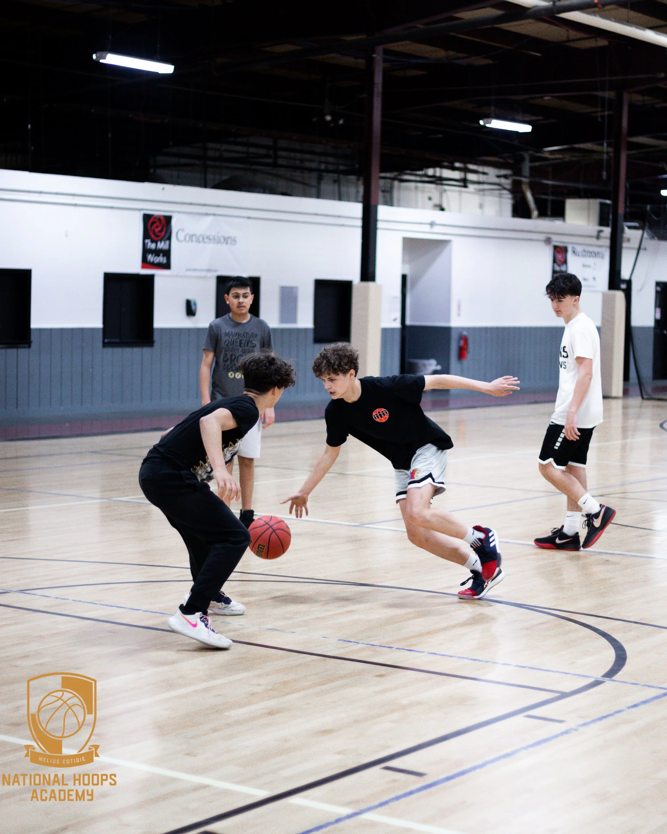 Four boys playing basketball on an indoor court, with one boy dribbling the ball while the others defend, and a logo for National Hoops Academy in the bottom left corner.