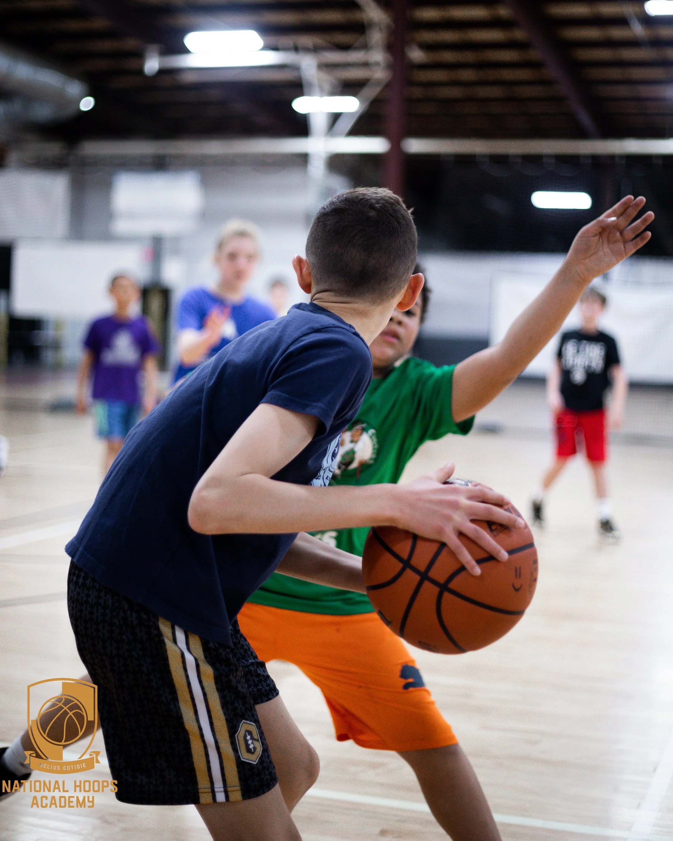 Children playing basketball on an indoor court, with two boys closely contesting the ball while other kids watch in the background.