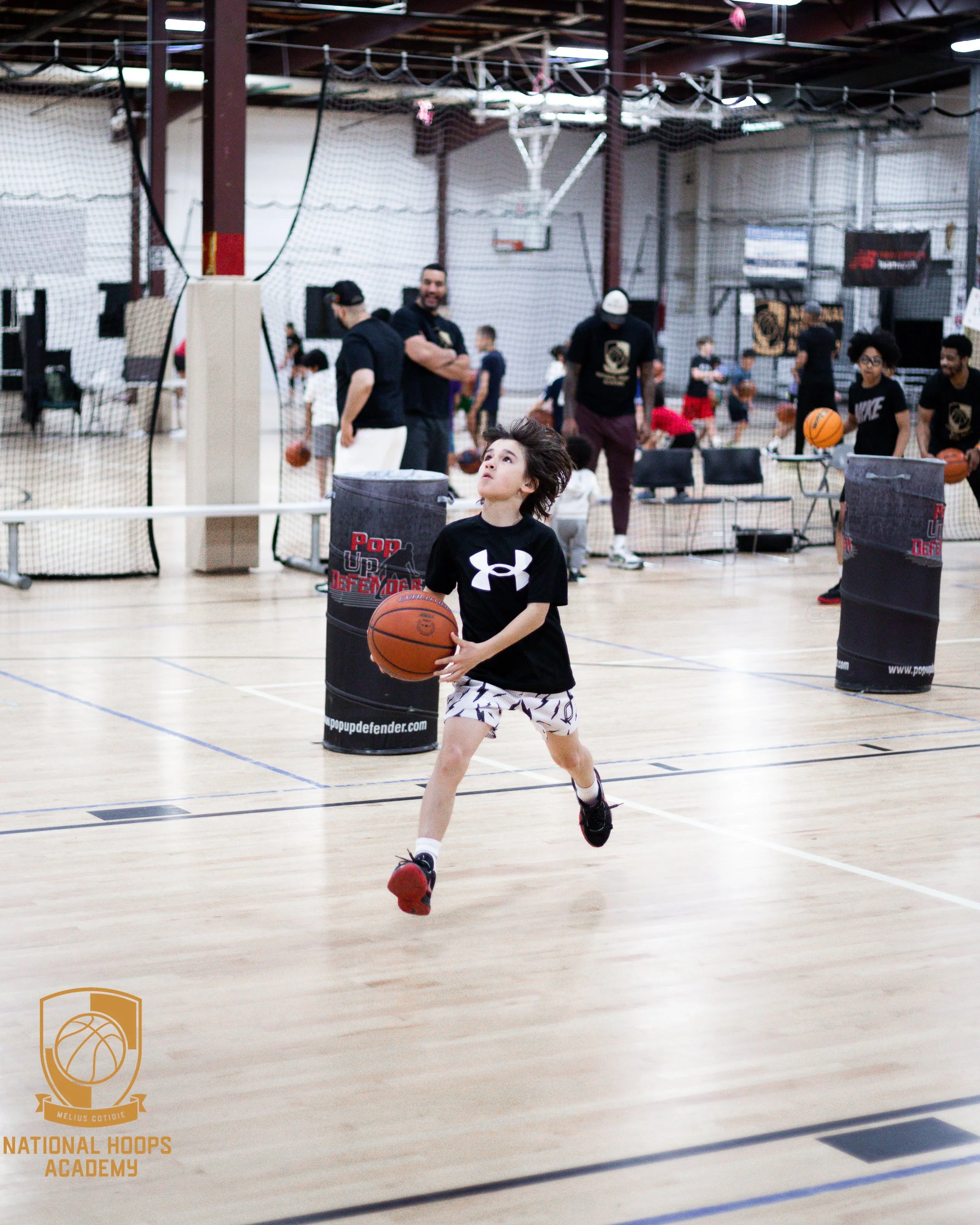 A young boy in athletic gear running with a basketball inside a gymnasium, with basketball training equipment and coaches visible in the background.