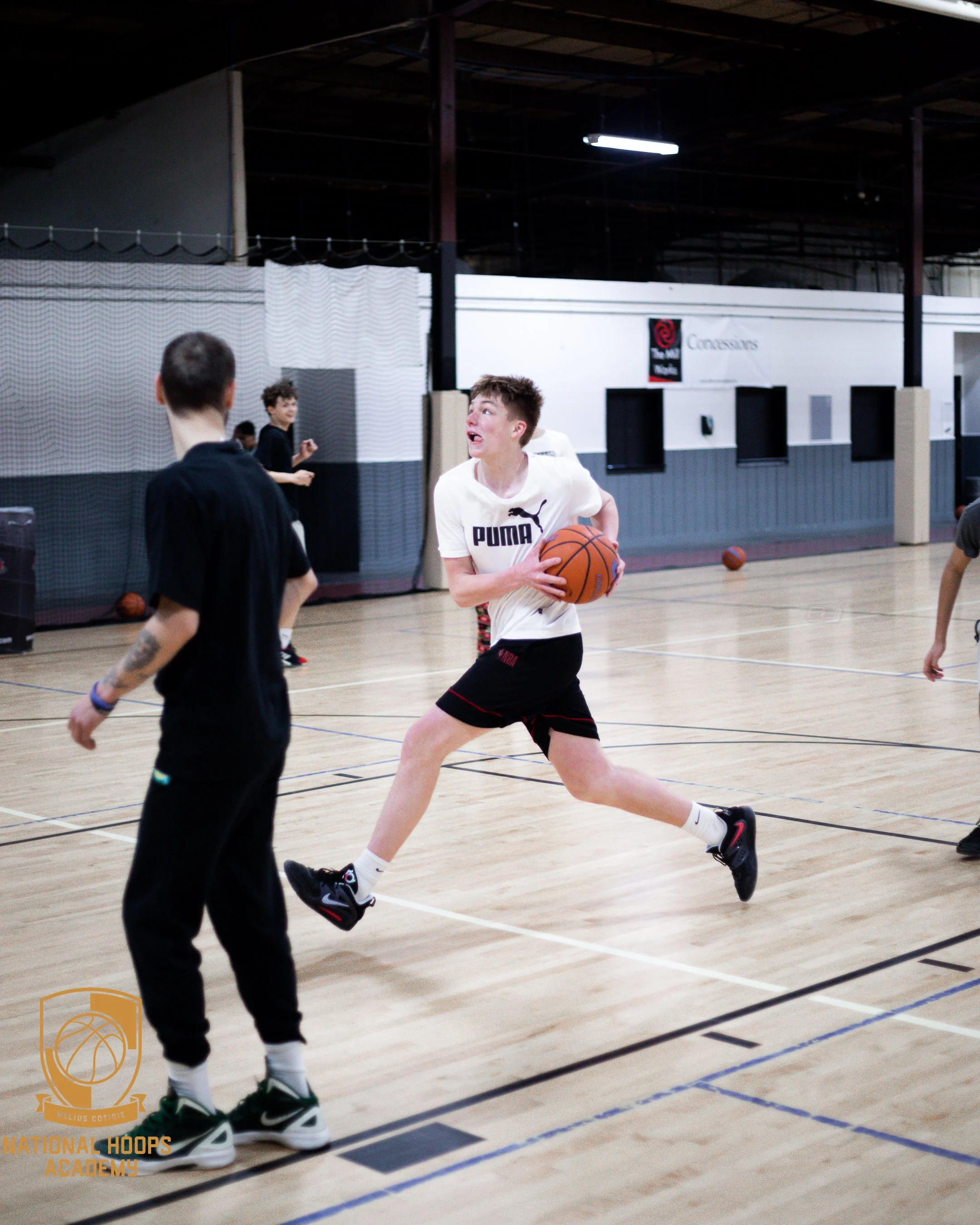 A boy in a white Puma shirt and black shorts dribbling a basketball on an indoor basketball court, with other kids in the background.