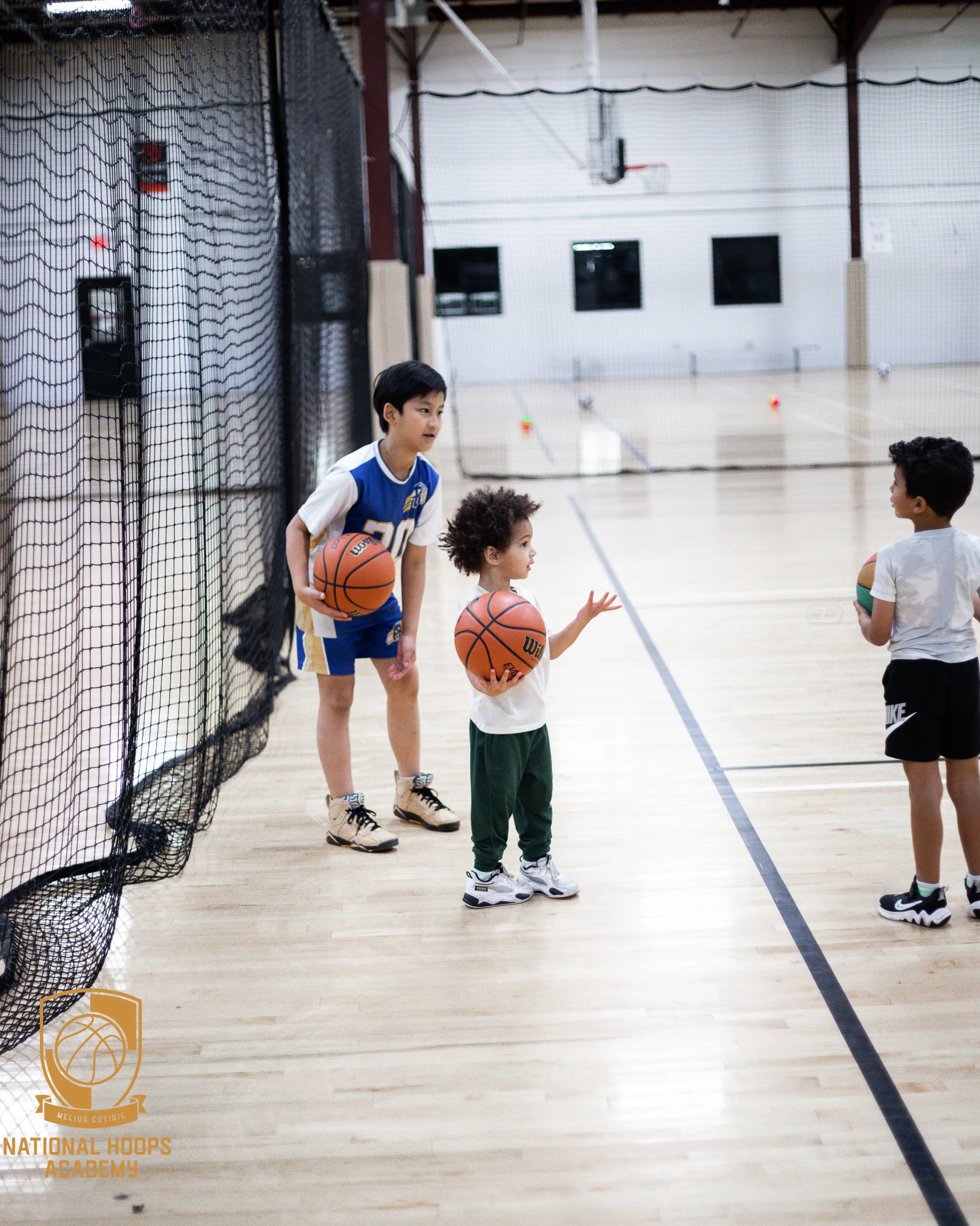 Three children standing on a basketball court inside a gym, holding basketballs, engaged in a basketball practice or lesson.