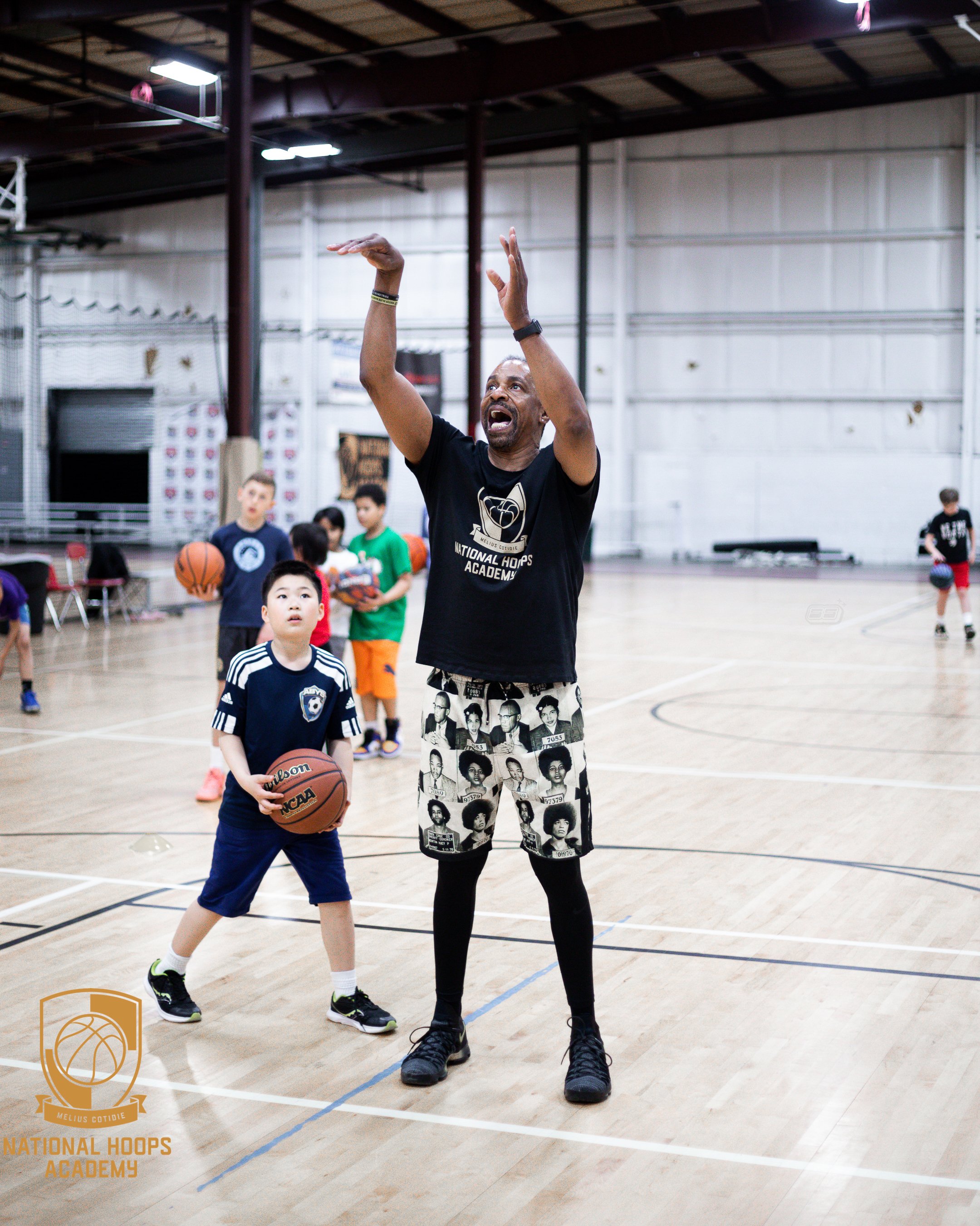 A basketball coach instructs young players during a practice session inside a gymnasium. The coach is actively demonstrating a technique, while children with basketballs listen. The gym has a high ceiling, hardwood floors, and sports equipment.