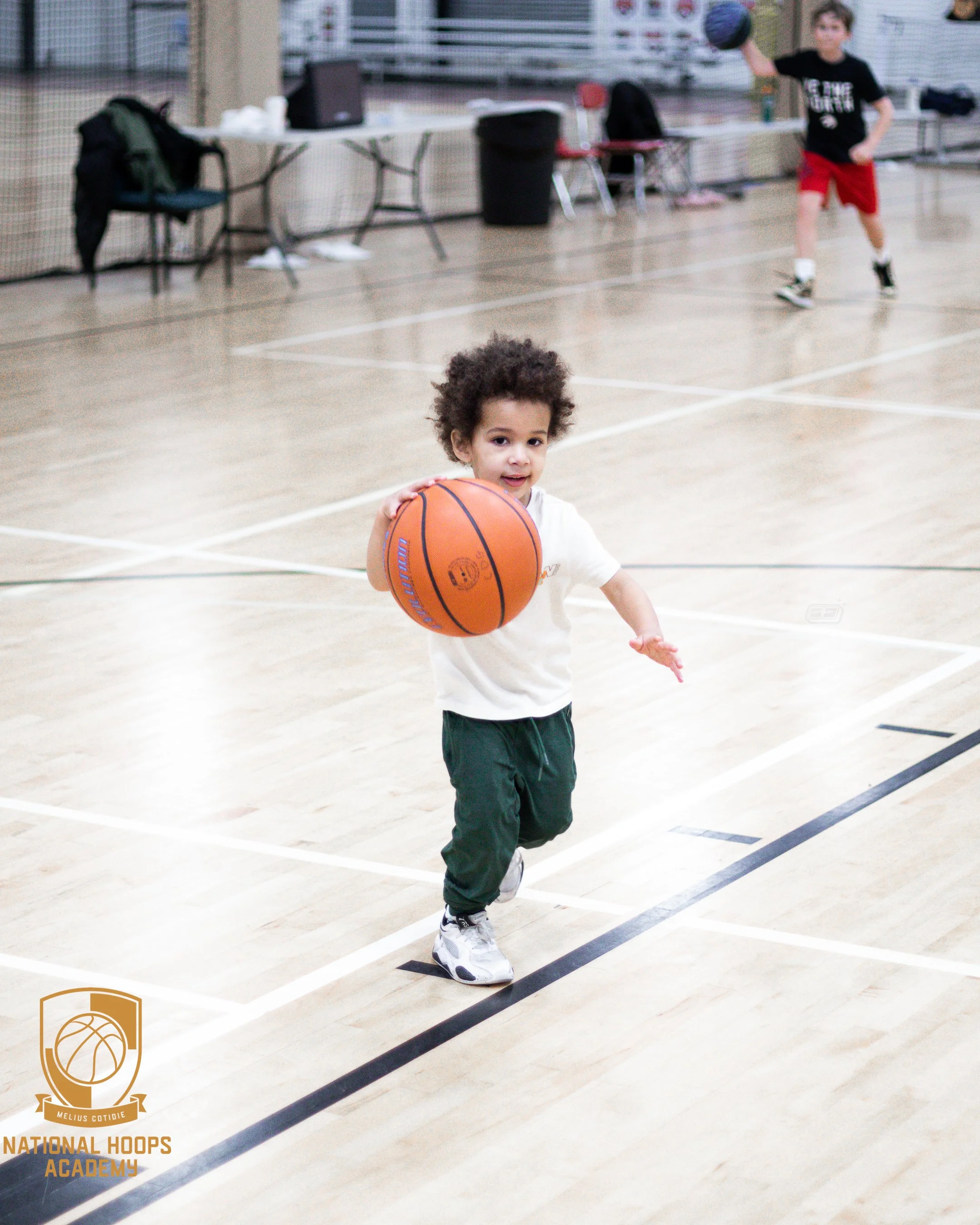 A young boy with curly hair wearing a white t-shirt, green pants, and white sneakers is running on an indoor basketball court while holding an orange basketball. In the background, a taller boy is playing with a basketball, and there are tables, chairs, and sports equipment around the court.