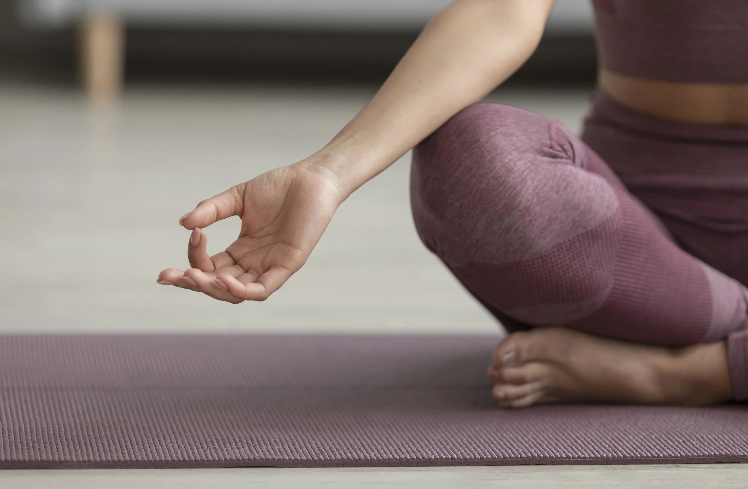 Close-up of a person in a yoga pose, wearing pinkish-purple workout clothes, with one hand on a yoga mat and the other hand resting on their foot, practicing meditation or yoga.