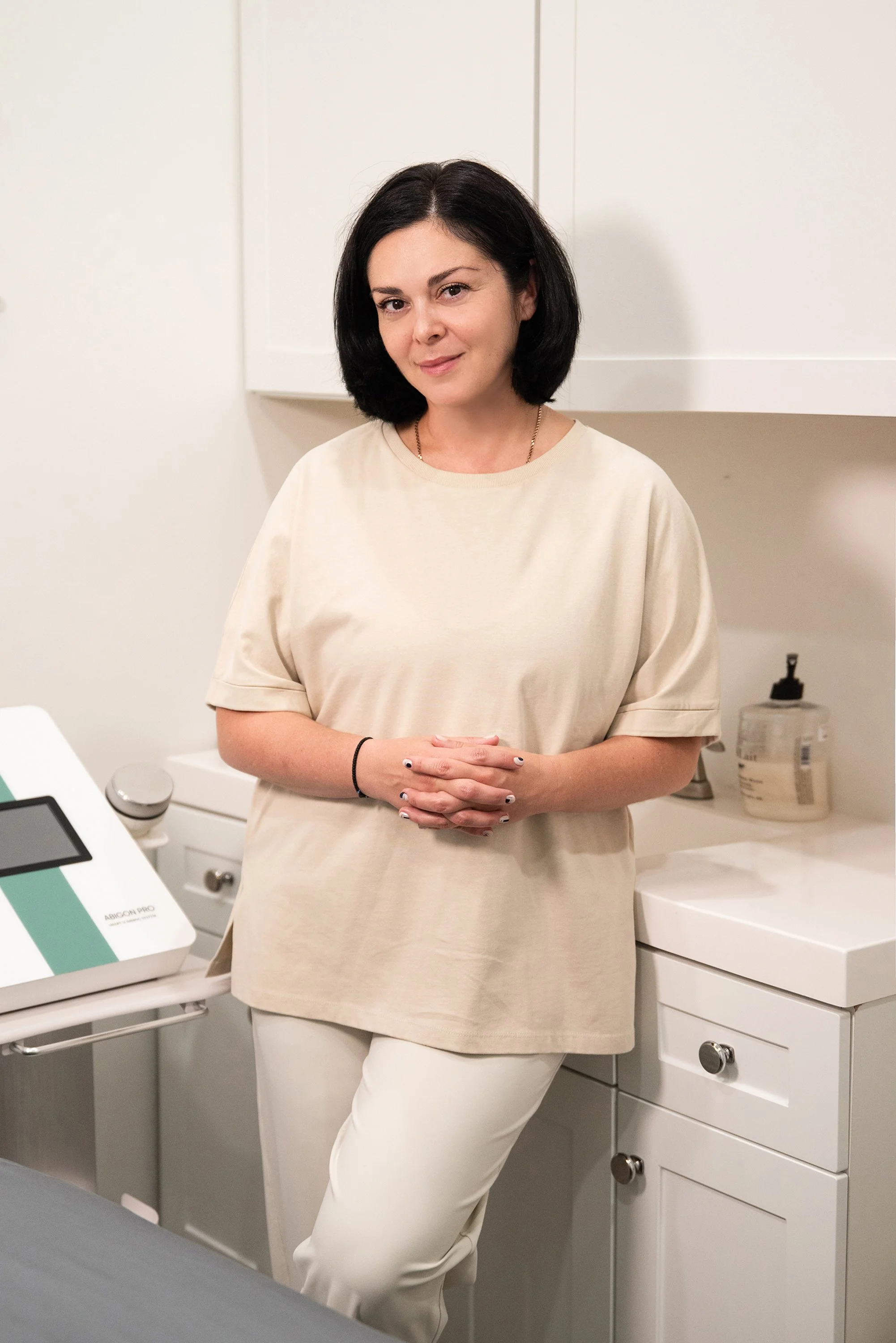 A woman with black hair wearing a beige T-shirt and white pants standing in a medical or clinical setting.