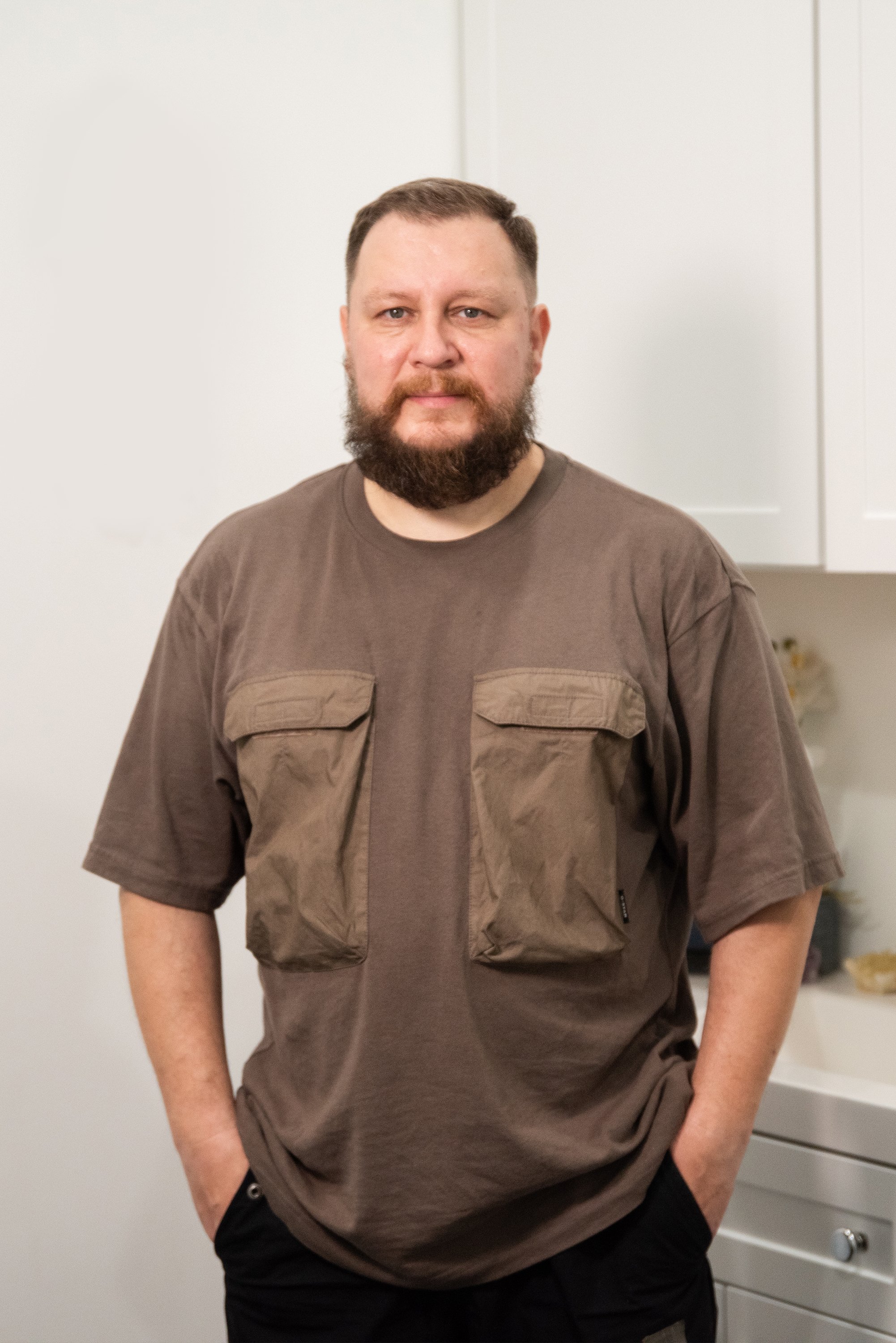 A man with a beard and short hair standing in a kitchen with white cabinets, wearing a brown T-shirt with two large front pockets and black pants, with his hands in his pockets.