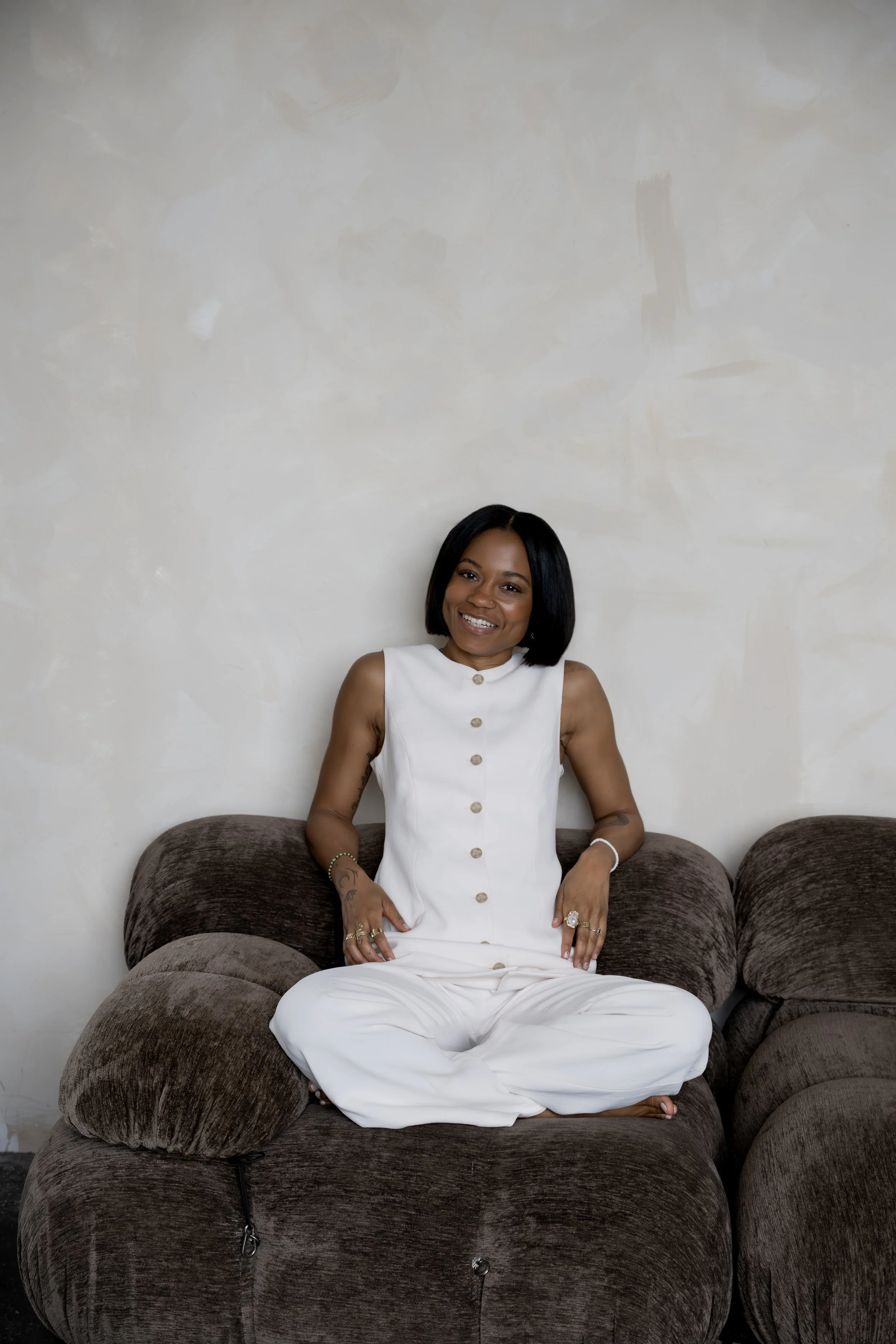 A woman with short black hair sitting on a dark brown plush couch, wearing a sleeveless white dress with buttons down the front, smiling and looking at the camera, with a plain off-white wall in the background.