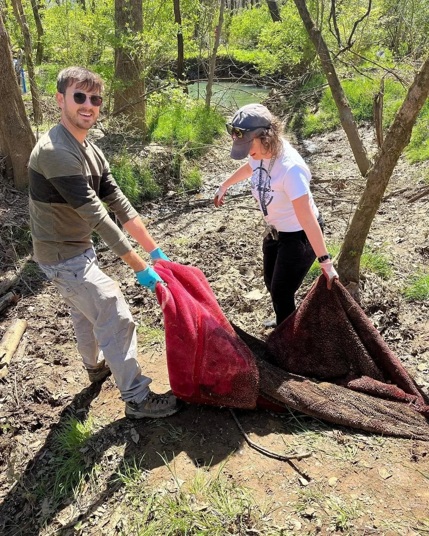 Two people in a wooded area are planting or removing a large, heavy, red and black object, possibly a piece of equipment or debris, near a creek.
