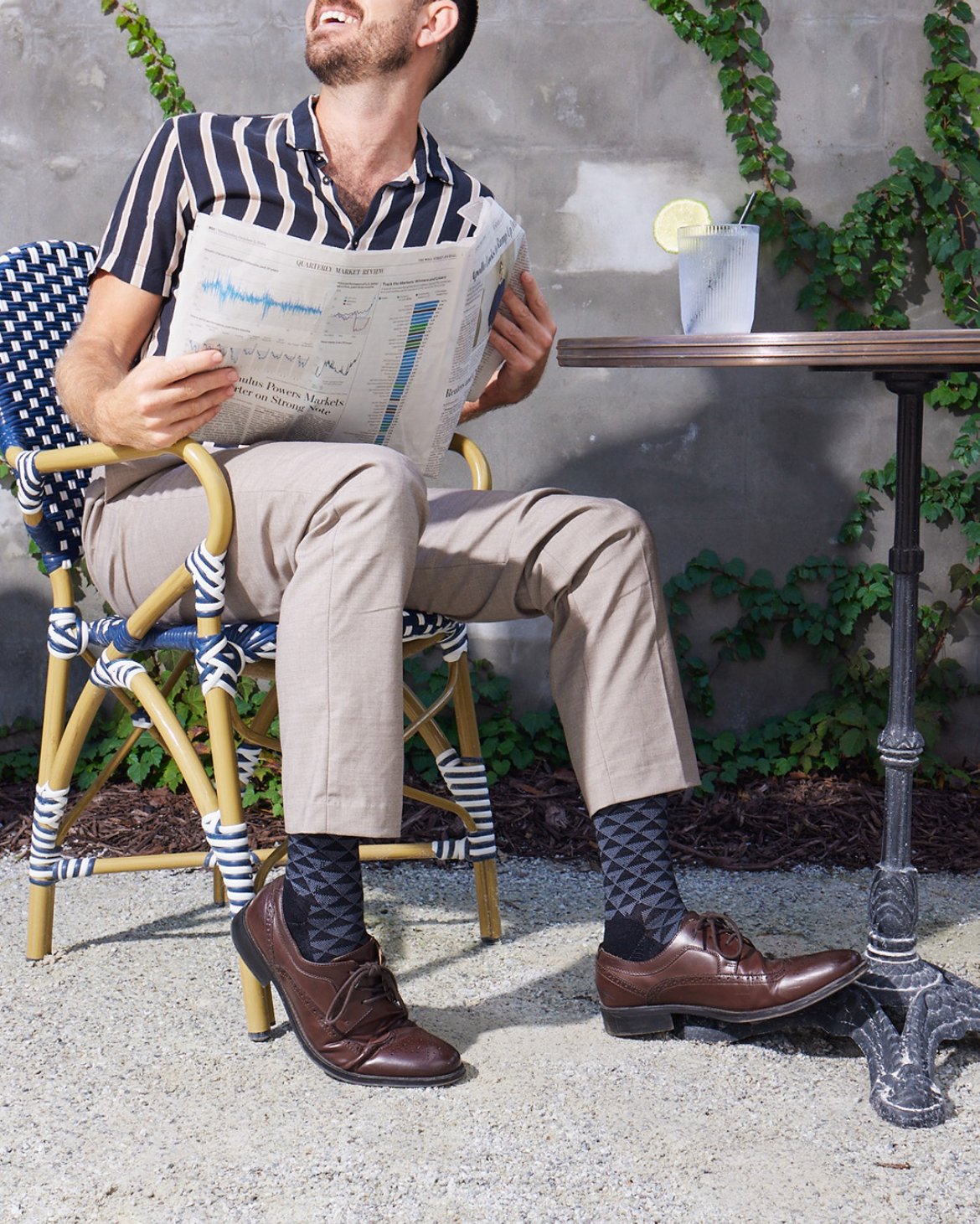 James sitting outside on a patterned chair, reading a newspaper, with a glass of water with a lemon slice on a small table beside him.