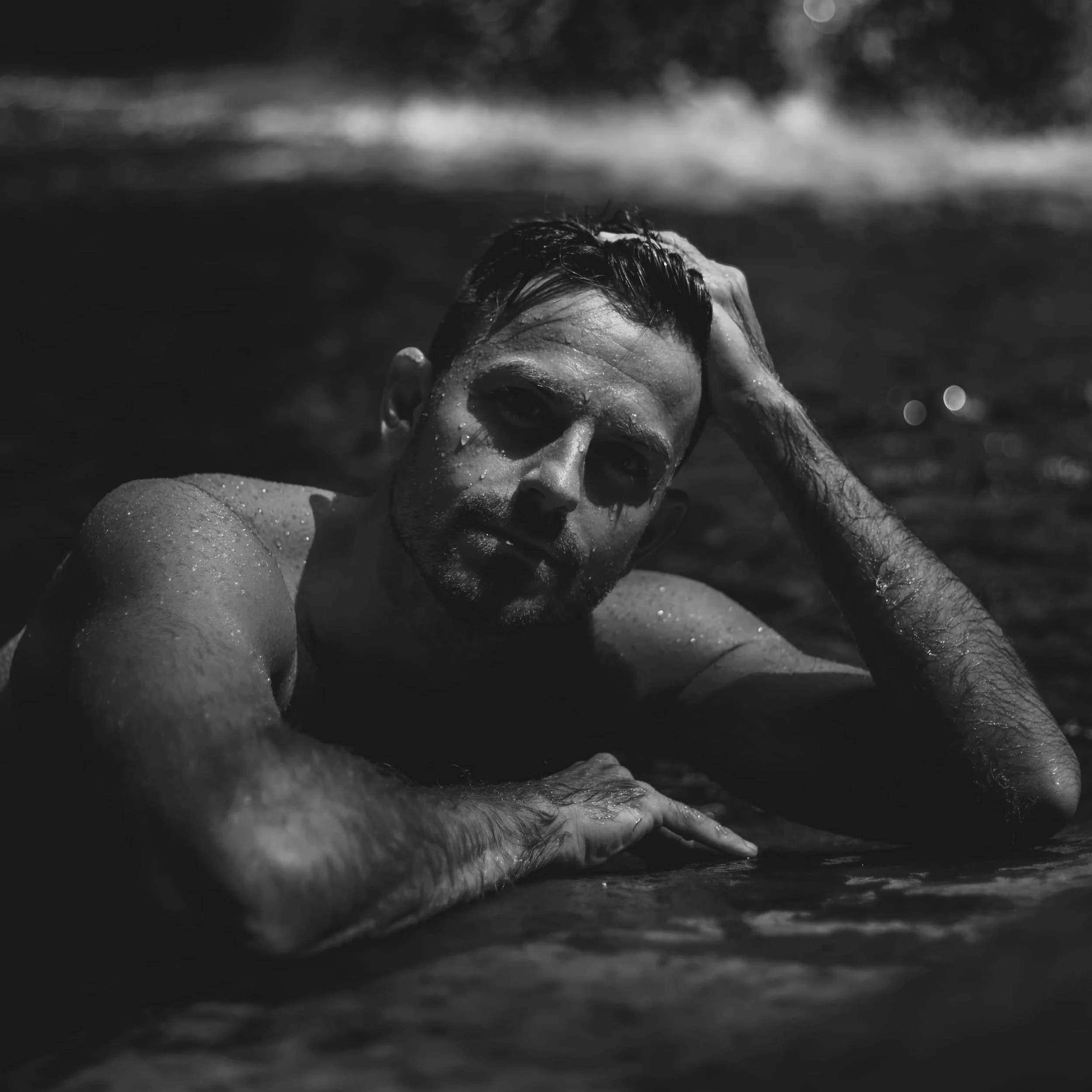 Black and white photo of James in a swimming hole, resting his head on his hand with water droplets on his skin, looking directly at the camera.