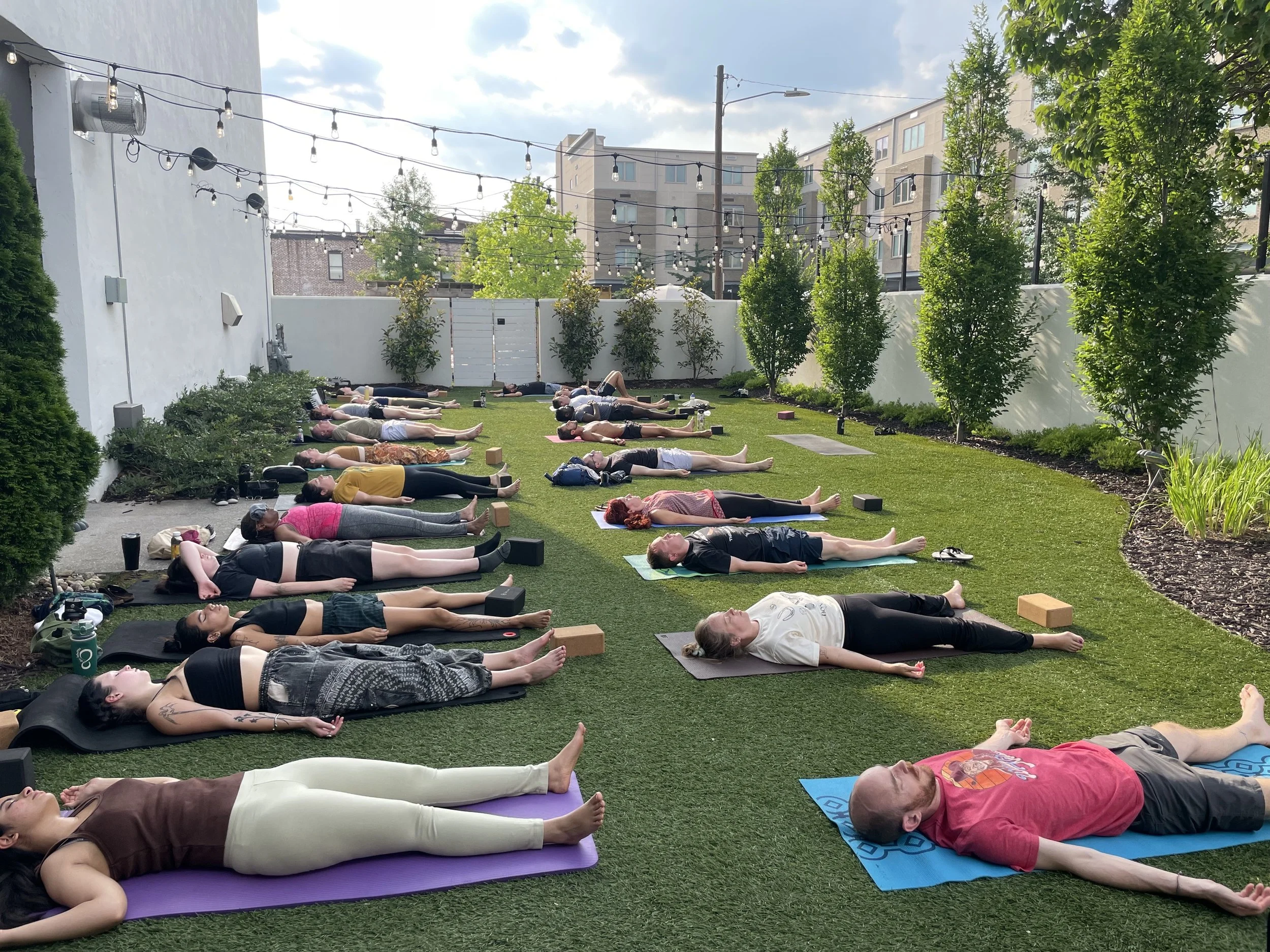 People participating in an outdoor yoga class on a grassy area surrounded by trees and a white fence, with buildings in the background and string lights overhead.