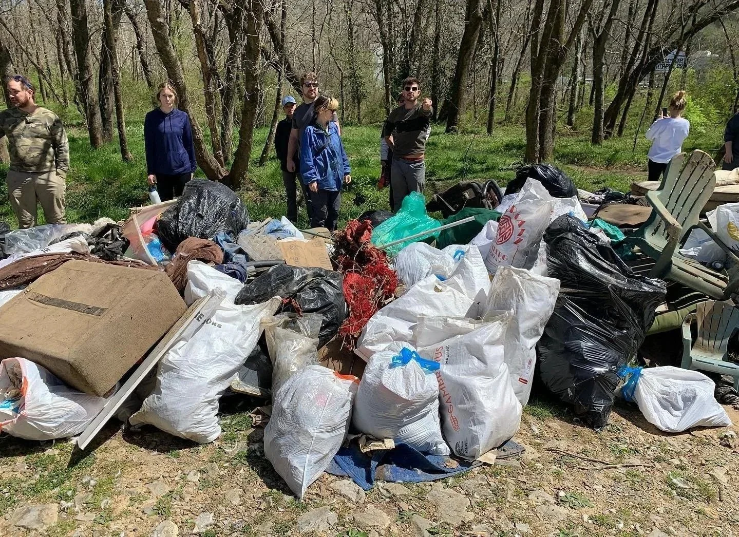 Group of people standing near a large pile of collected trash and garbage bags in a wooded area during a cleanup event.