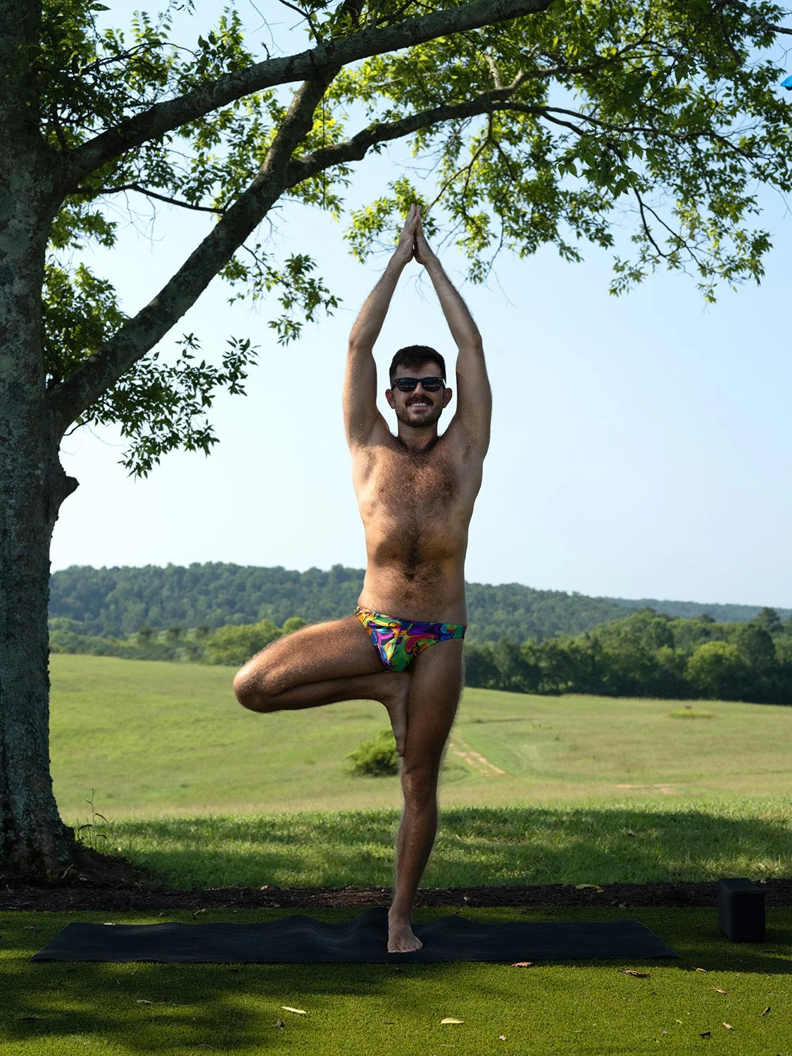 James with a beard and sunglasses doing a tree yoga pose outdoors on a yoga mat, wearing colorful swim trunks.
