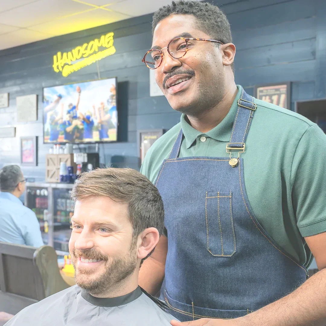 James getting a haircut in a barbershop, smiling and sitting in a barber chair, while the barber, wearing glasses and an apron, stands beside him smiling. The background features a television and a neon sign that says 'Handsome'.