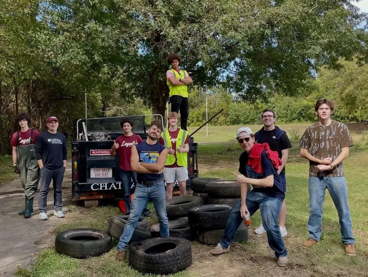 A group of ten young men posing outdoors near a pickup truck with tires on the ground. Some are standing and one is sitting on a tire, all smiling or making gestures. The setting is a grassy area with trees in the background.