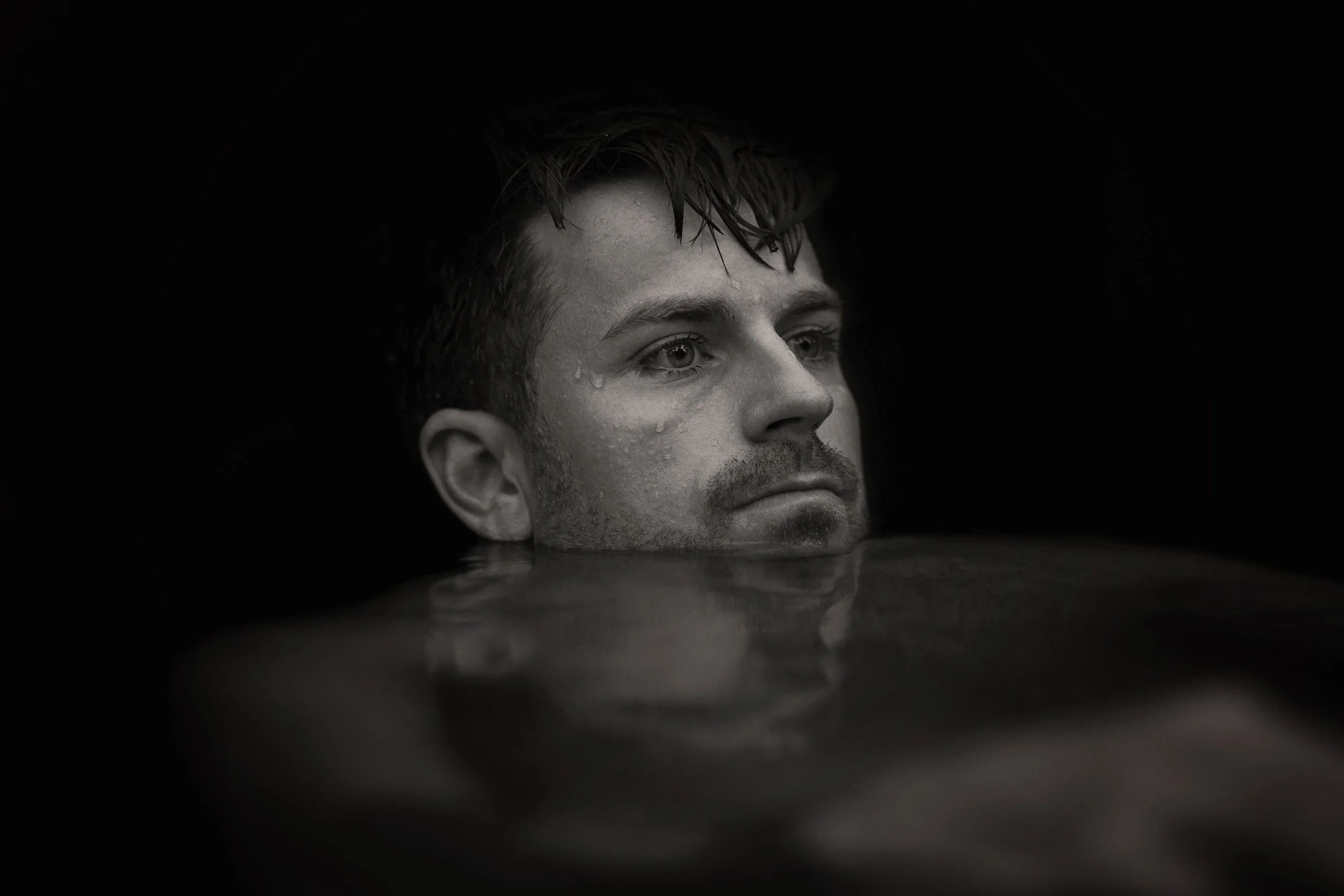 A black and white close-up of James's face partially submerged in water, with water droplets on his cheek and a dark background.