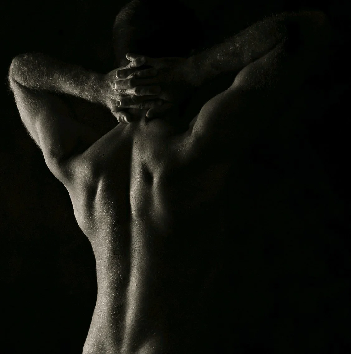 A black and white photograph of James's back and shoulders with hands clasped behind the head, highlighting the muscular contours and skin texture.