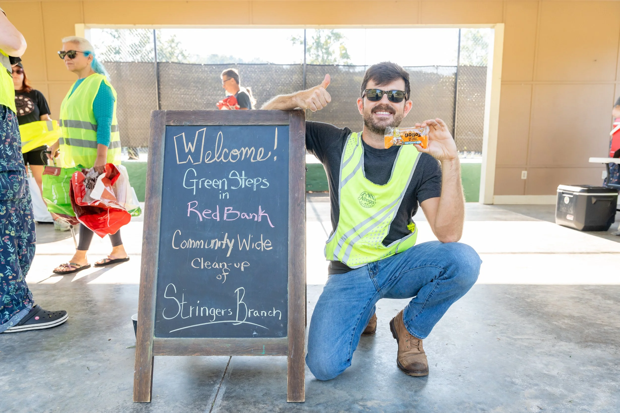 James kneeling beside a welcome sign at a community cleanup event, holding a snack and giving a thumbs-up.