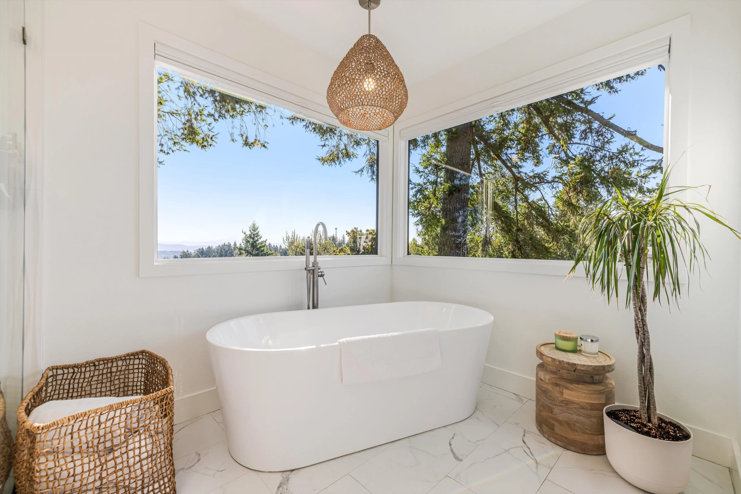 Bright bathroom with a white bathtub by large windows showing trees and a blue sky, with a woven basket, a potted plant, and candles.