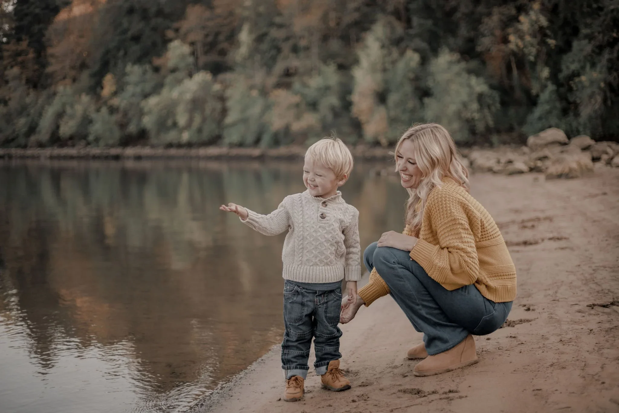 A woman and a young boy smile and play by a riverbank during autumn, with trees in the background.