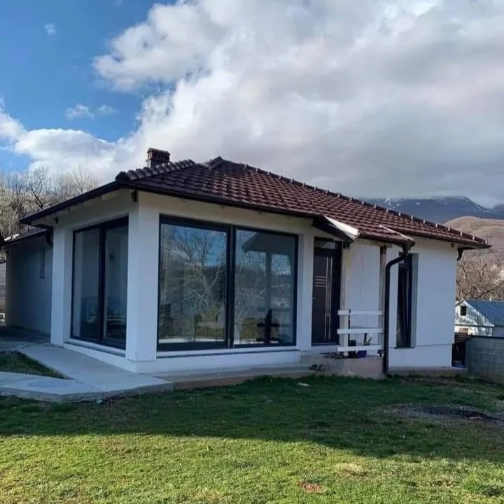 A house with large glass windows, a sloped roof with red tiles, and a small white bench outside, surrounded by grass and mountains in the background.