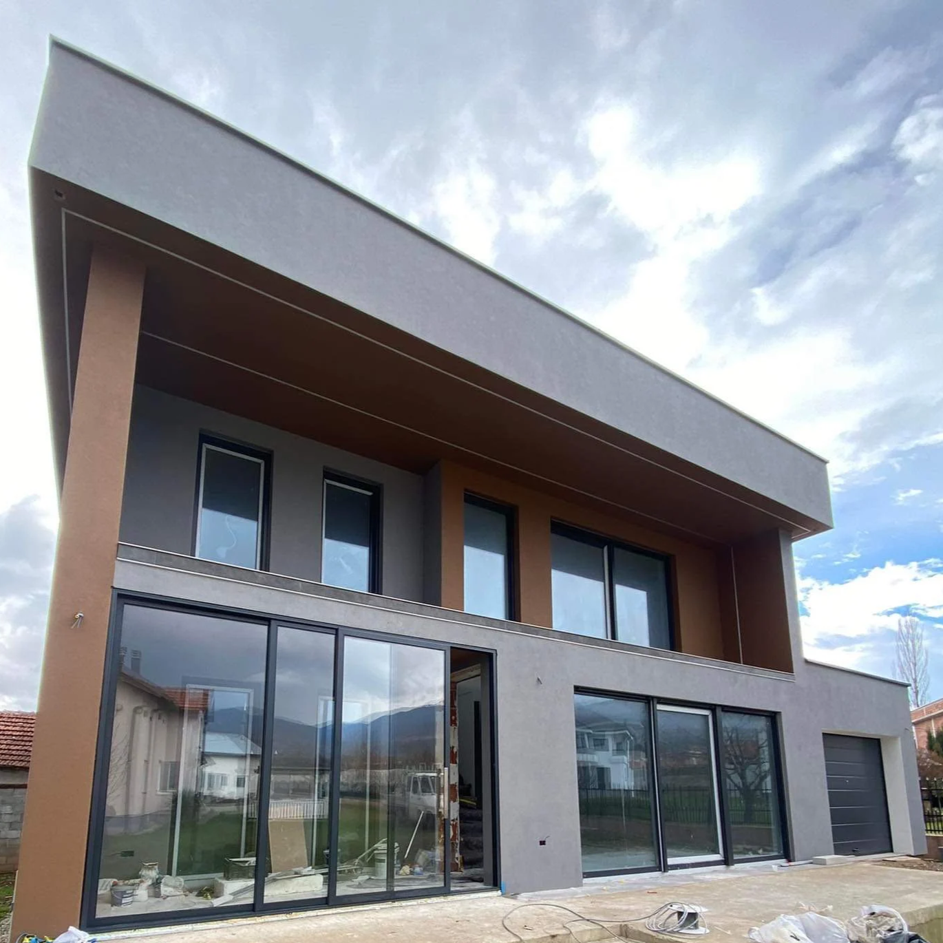 Modern two-story house under construction with large glass sliding doors and windows, and a flat roof, set against a partly cloudy sky.