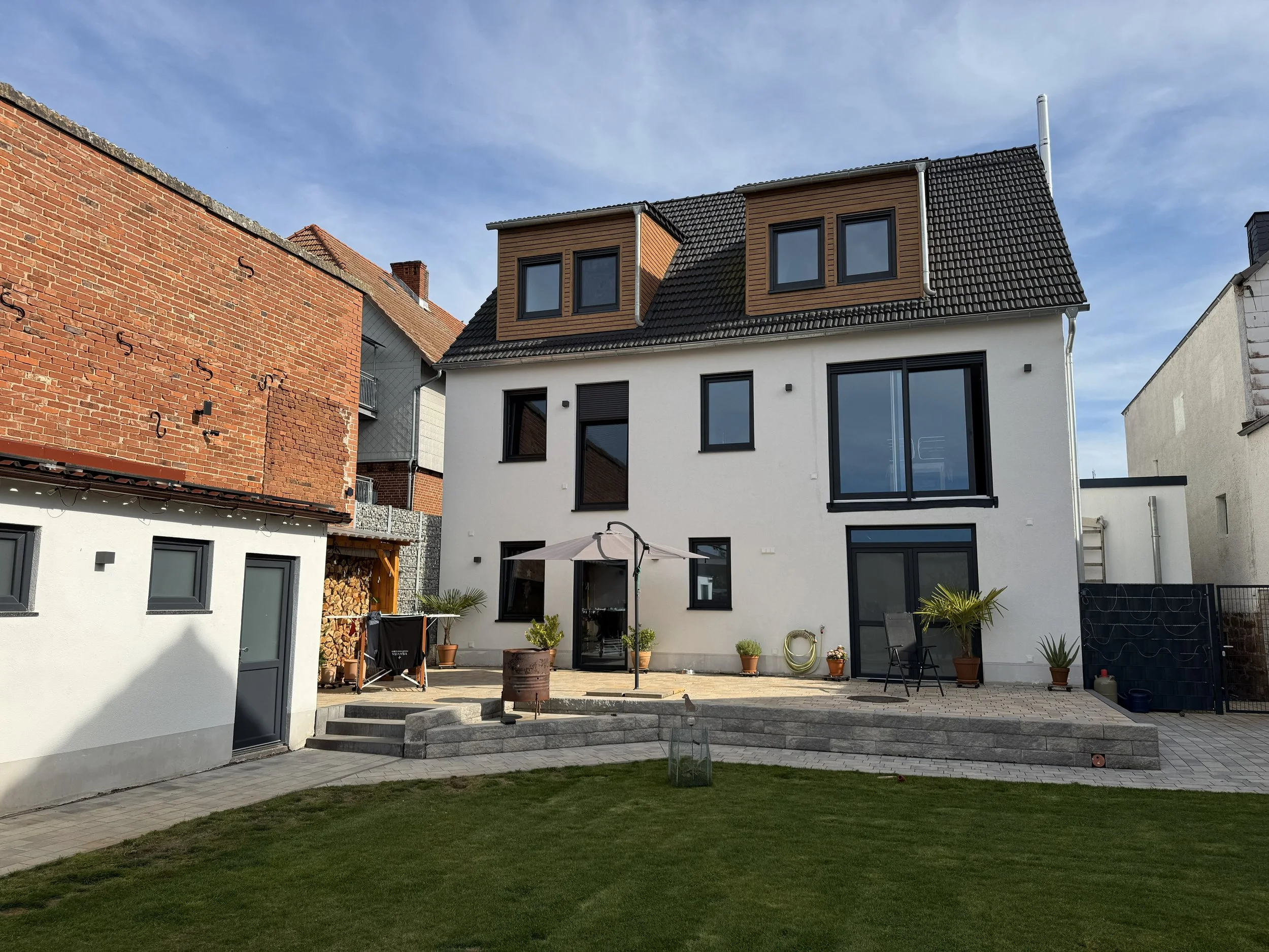 Modern white house with large windows, a patio with potted plants, chairs, and an umbrella, surrounded by a well-maintained lawn and neighboring buildings, under a partly cloudy sky.