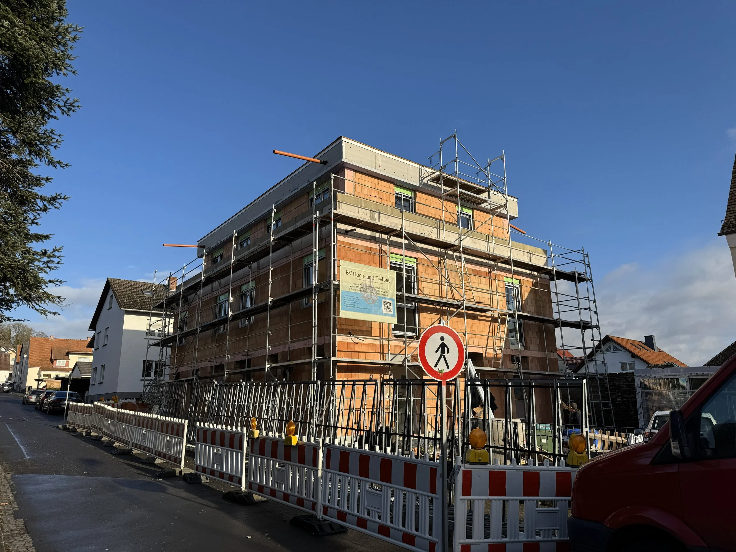 A multi-story building under construction with scaffolding around it, located on a residential street, with a no pedestrian sign and construction barriers in the foreground.