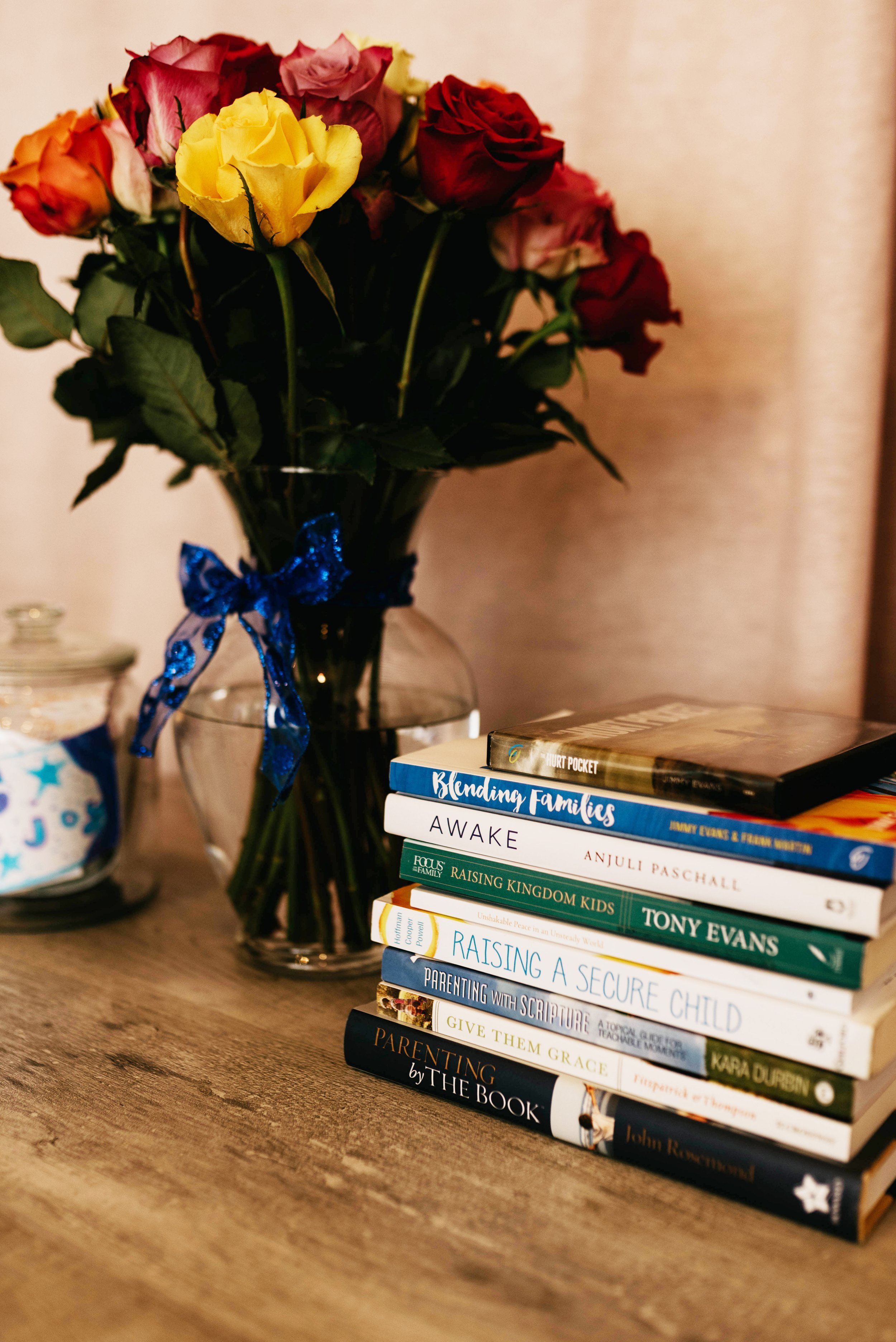 A bouquet of multicolored roses in a glass vase with a blue ribbon tied around it, placed on a wooden surface. Next to the bouquet is a stack of books with titles related to parenting and family topics.