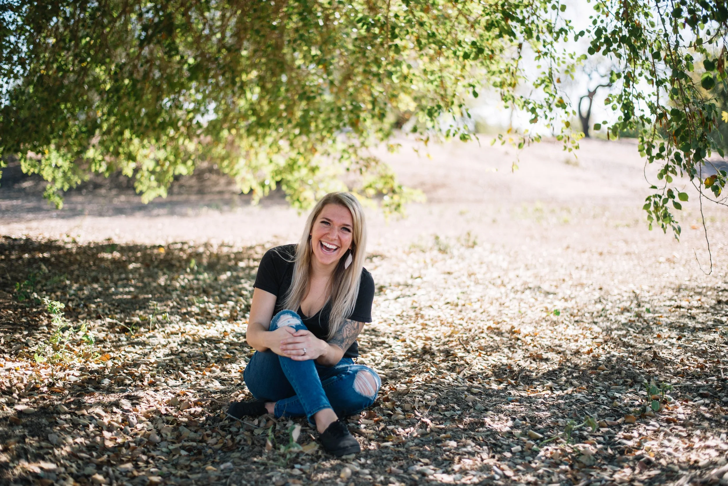A woman sitting on the ground under a large tree, smiling and laughing, surrounded by fallen leaves in a park or outdoor setting.