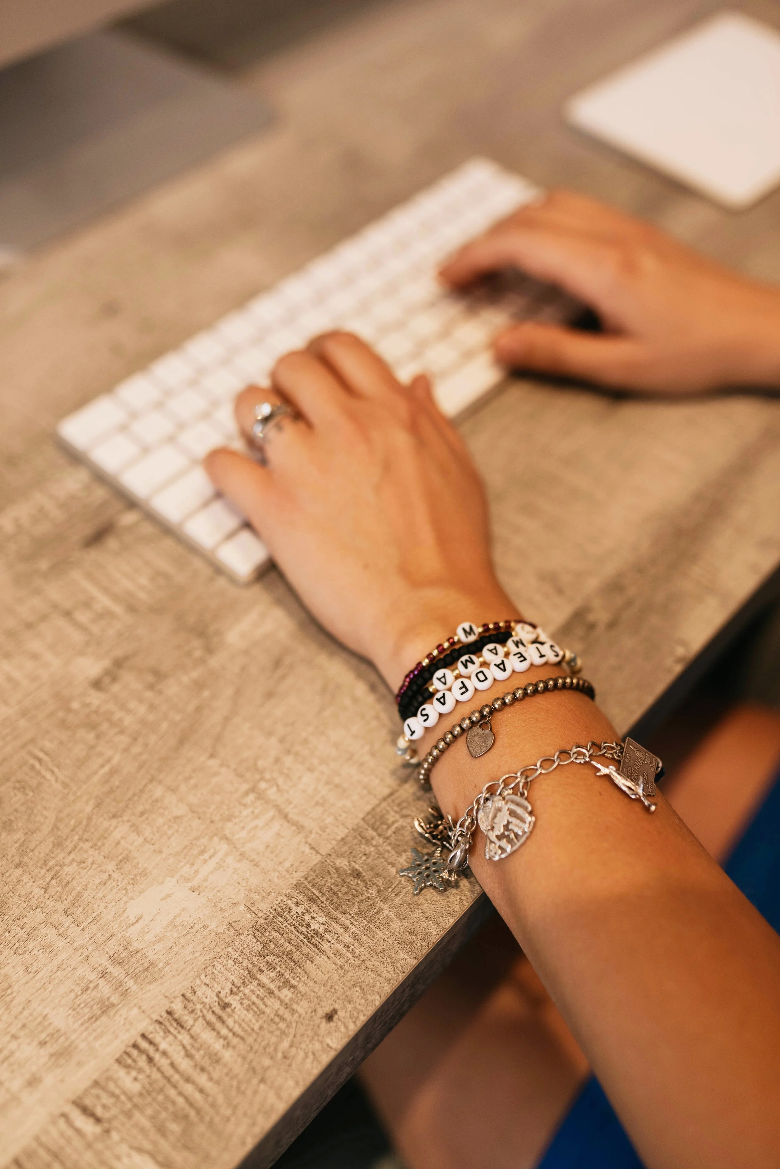 A person's hand typing on a white computer keyboard on a rustic wooden desk, wearing several bracelets including one with letter beads spelling 'M A D E R A S T'