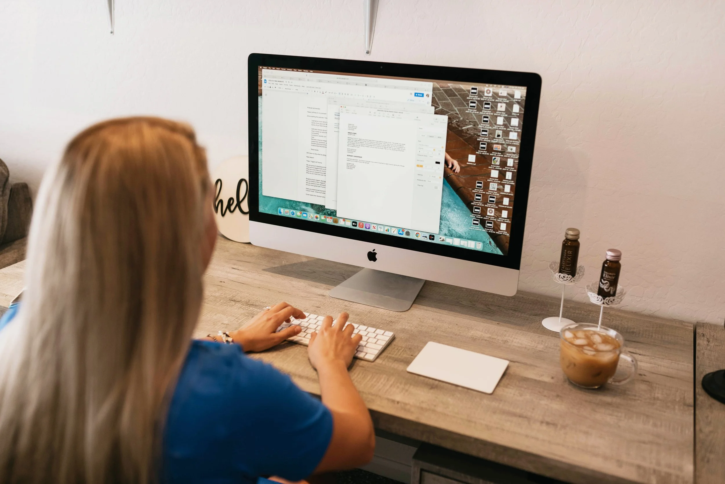 A woman with blonde hair working on an iMac computer at a wooden desk with a cup of iced coffee and two small bottles of flavored syrup nearby.