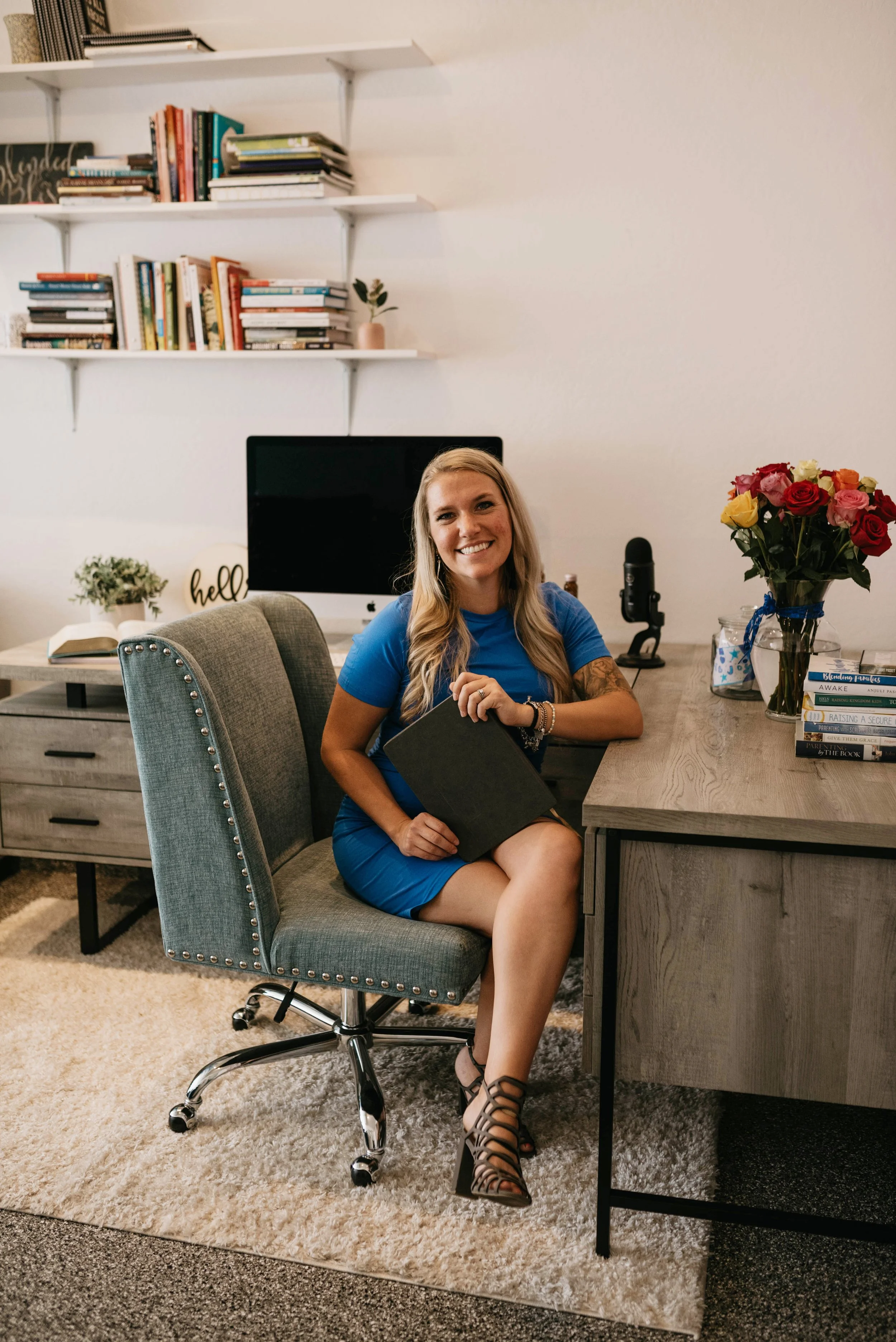 A woman sitting at a desk in a home office, holding a black notebook and smiling at the camera, with a computer, a microphone, a vase of roses, and a stack of books on the desk, and bookshelves with books and a small plant in the background.