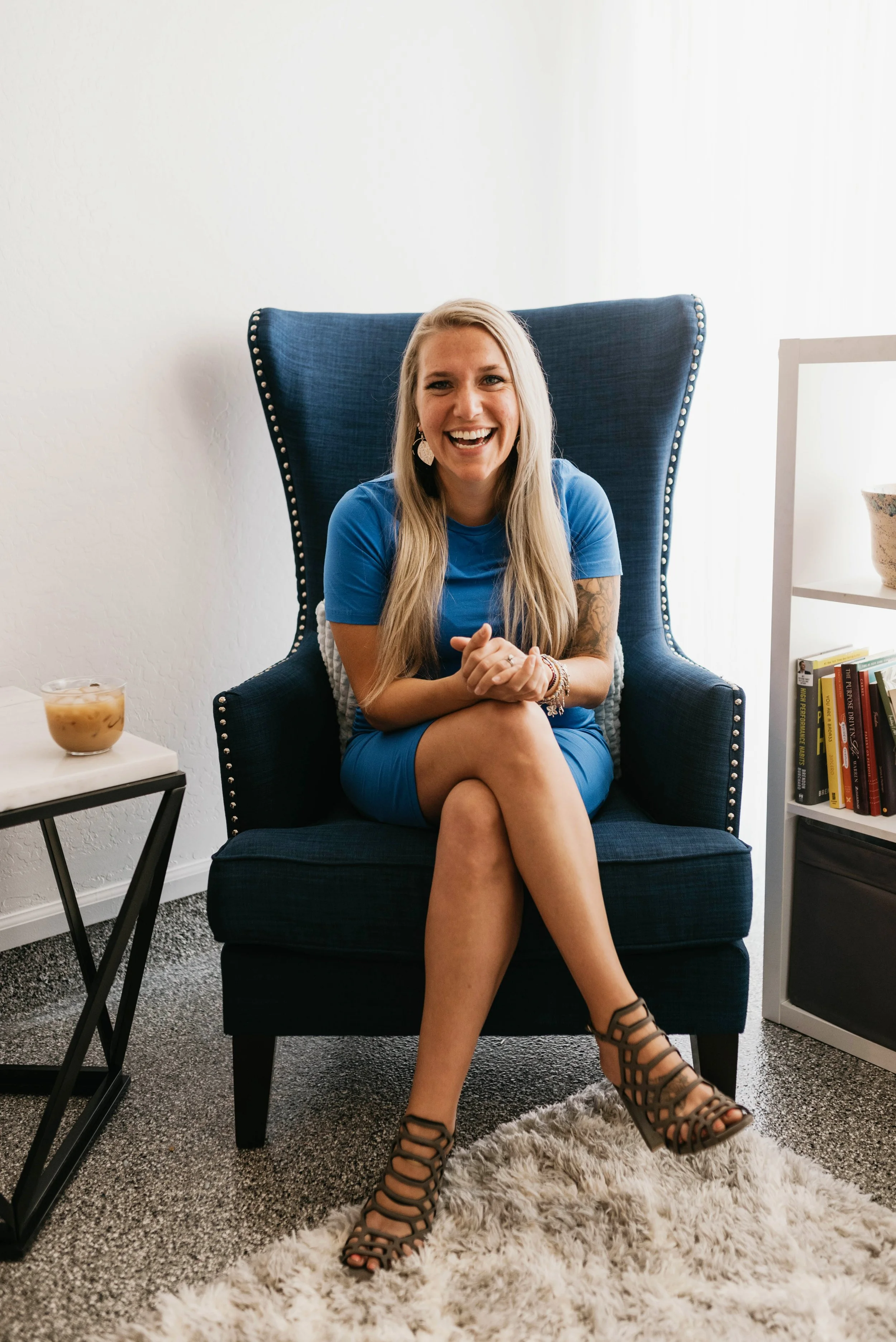 Woman with long blonde hair, smiling, sitting on a dark blue high-back chair in a living room, wearing a blue dress and black caged heels, with a cream-colored rug, a small table with a bowl, and a white bookshelf with books and decorative items.