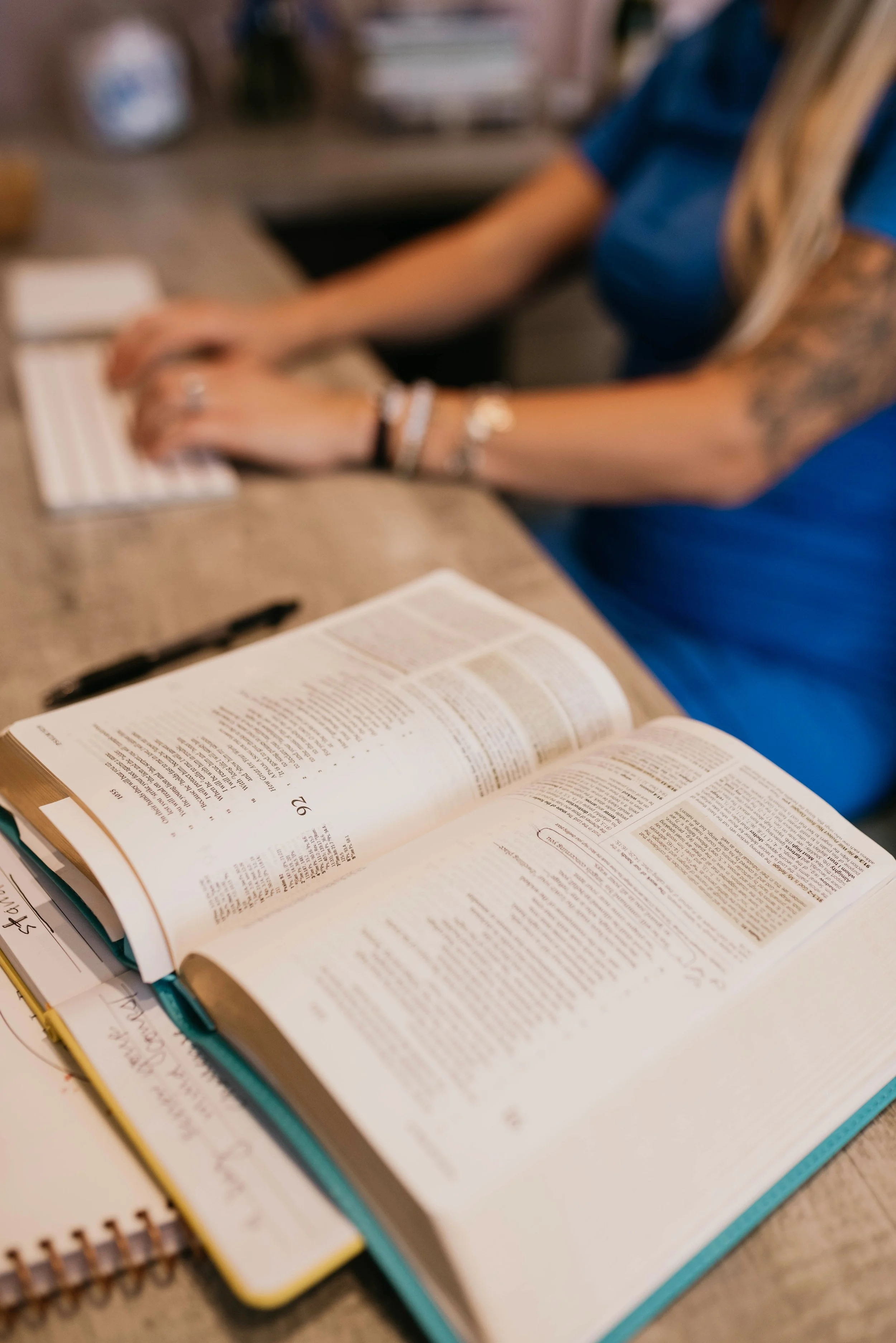 Open Bible and notebooks on a wooden table, with a woman wearing a blue shirt sitting nearby, in a cozy indoor setting.