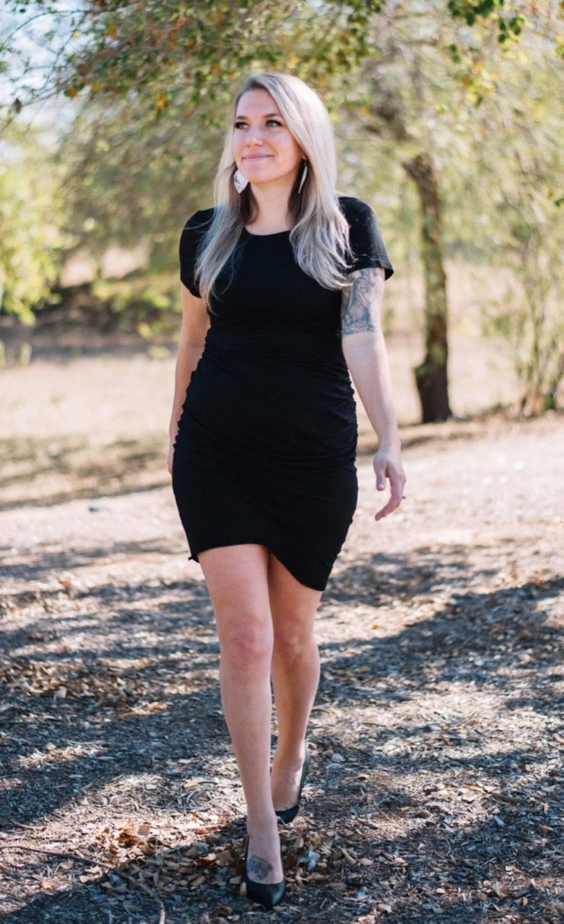 A woman with long, light blonde hair, wearing a black dress and black heels, walking outdoors on a dirt path with trees in the background.