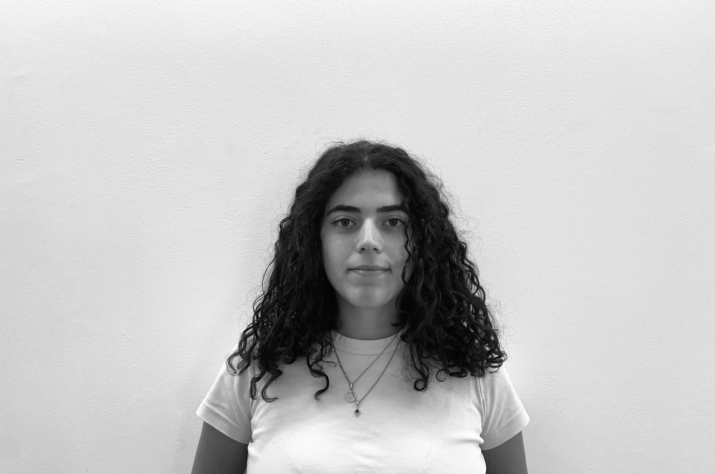 A young woman with curly hair and a neutral expression poses in front of a plain white wall, wearing a white t-shirt and layered necklaces.
