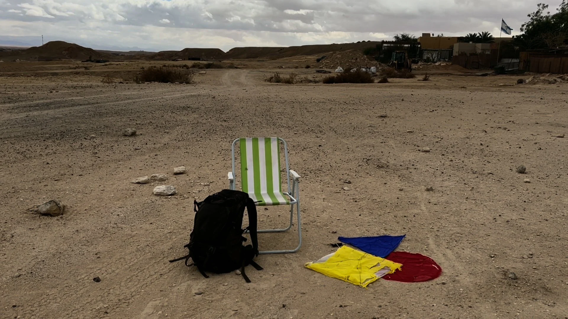 Empty desert landscape with a green and white striped chair, a backpack, and multicolored flags or fabric pieces on the ground in the foreground. Distant hills and some buildings are visible under a cloudy sky.