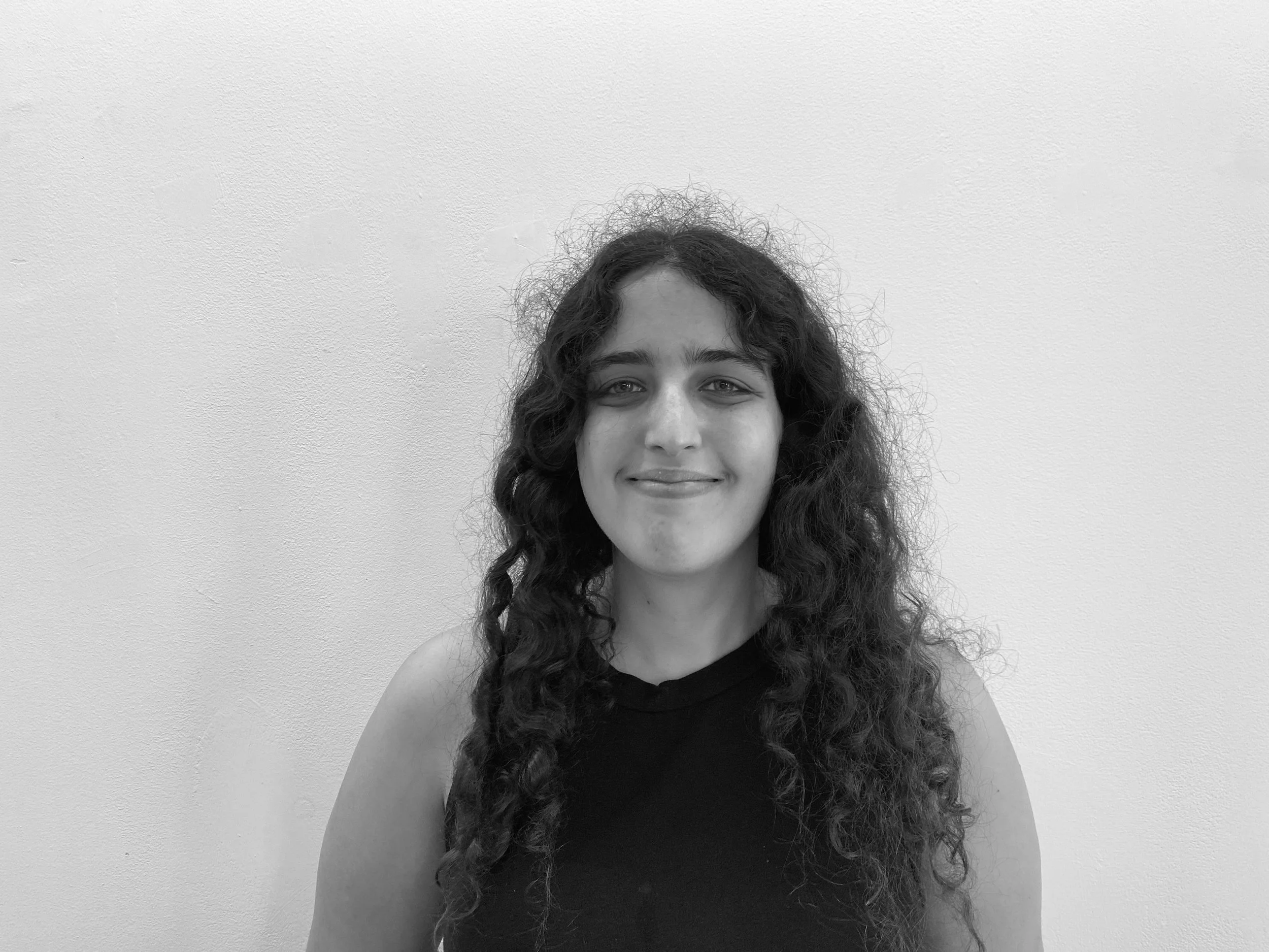 A young woman with curly hair smiling, wearing a sleeveless black top, against a plain white wall.