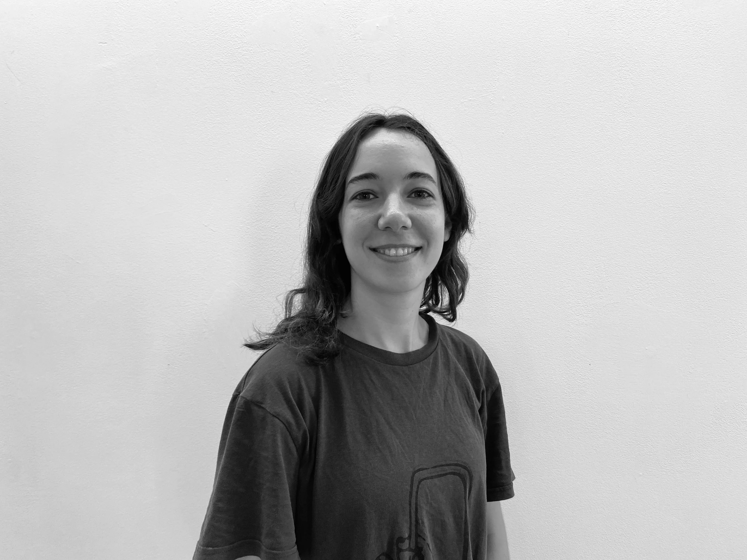 Black and white photo of a young woman with shoulder-length hair, smiling, standing against a plain wall.