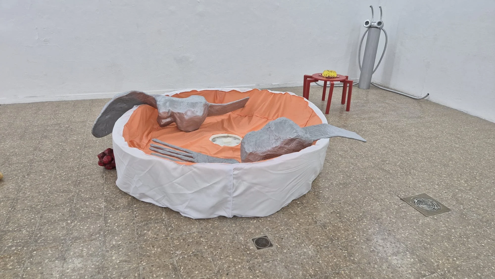 Art installation of a large paper bowl with sculpted gray limbs and a skull, set on a tiled floor in a gallery space with a white wall and a small red table holding a yellow object in the background.
