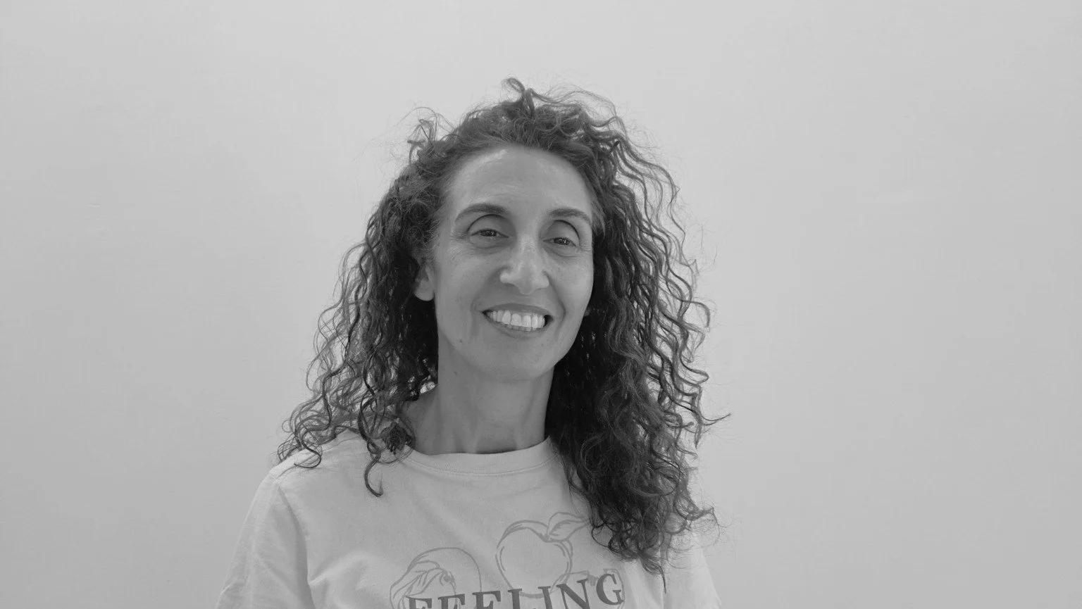 Black and white photo of a woman with curly hair smiling, wearing a t-shirt with text and apple illustrations, standing against a plain wall.