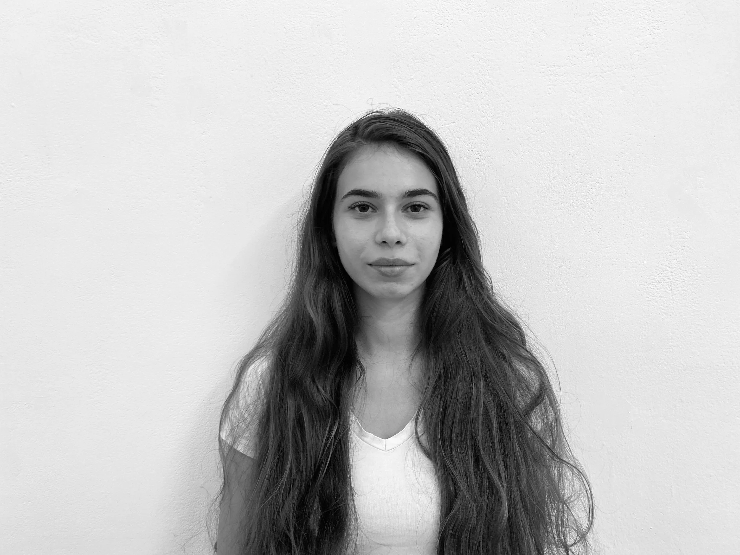 Black and white photograph of a young woman with long wavy hair, wearing a white top, standing against a plain light-colored wall.