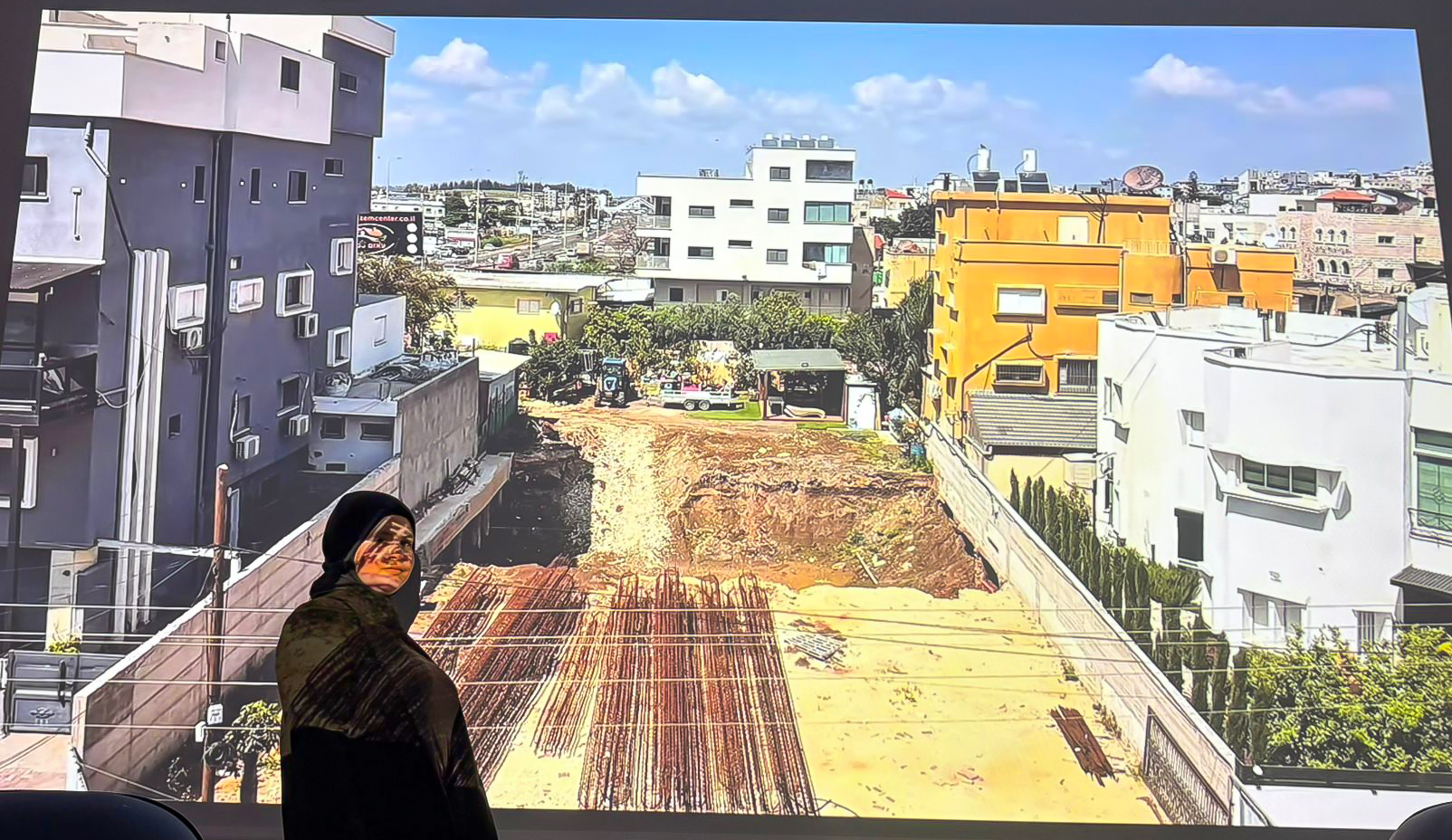 A woman wearing a black hijab stands in front of a large outdoor screen showing an image of a cityscape with colorful buildings, trees, and a construction site.