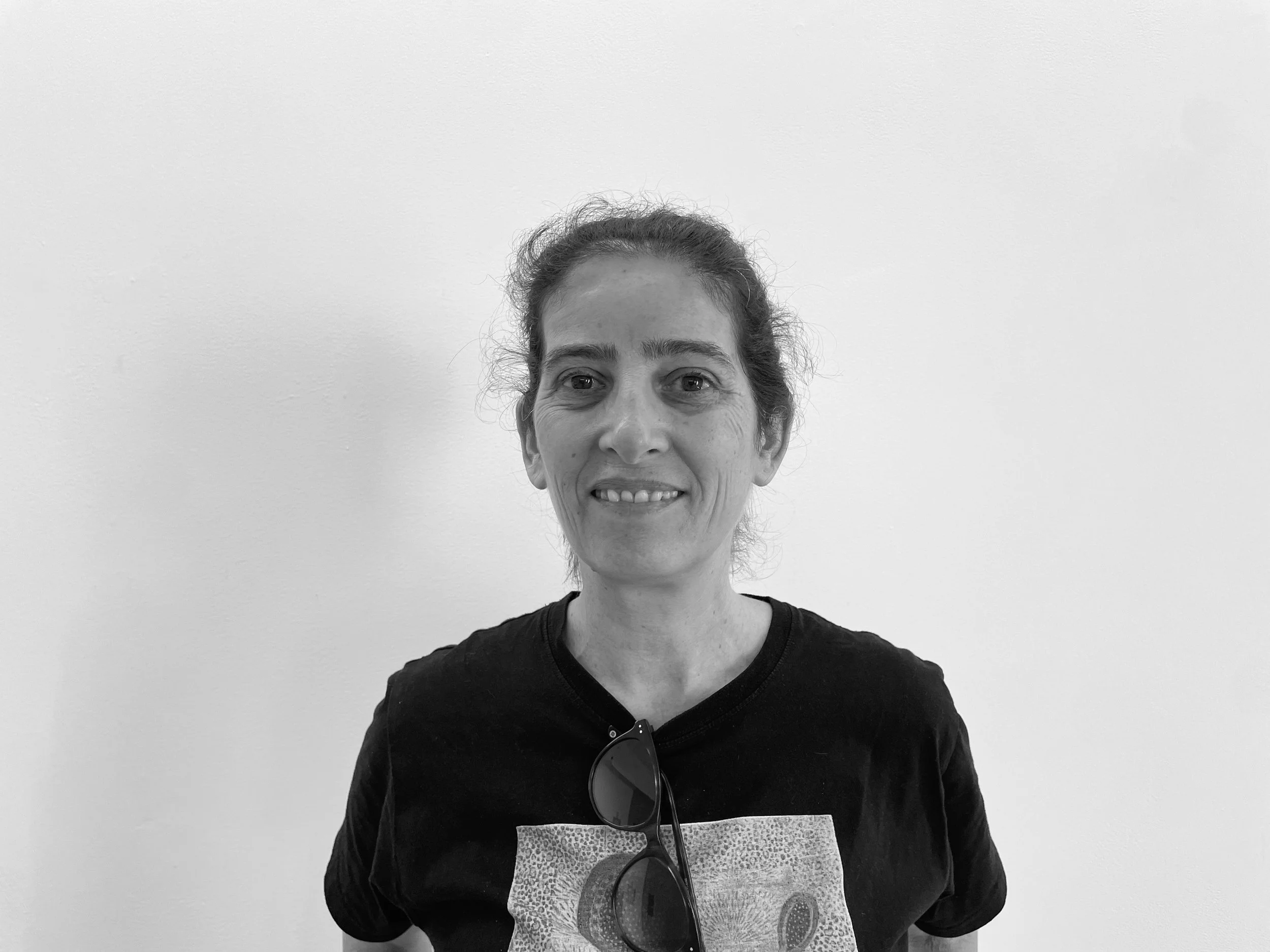 A woman with curly hair pulled back, smiling, wearing a black shirt and sunglasses hanging from the neck, standing against a plain white wall.